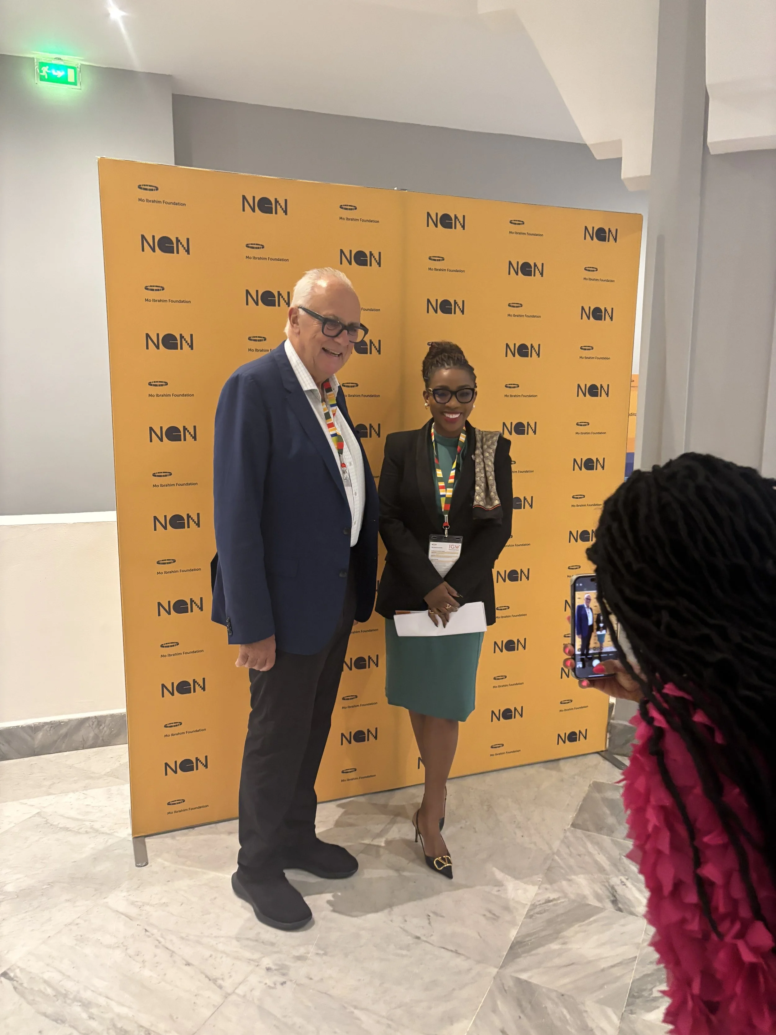 Two professionally dressed people standing in front of an orange backdrop with the NgN and Mo Ibrahim Foundation logos, smiling for a photo at an event. A woman in a teal dress and high heels is holding a white notepad, while a man in a suit and glasses is standing beside her. Another person with braided hair and a red textured outfit is taking a picture of them.