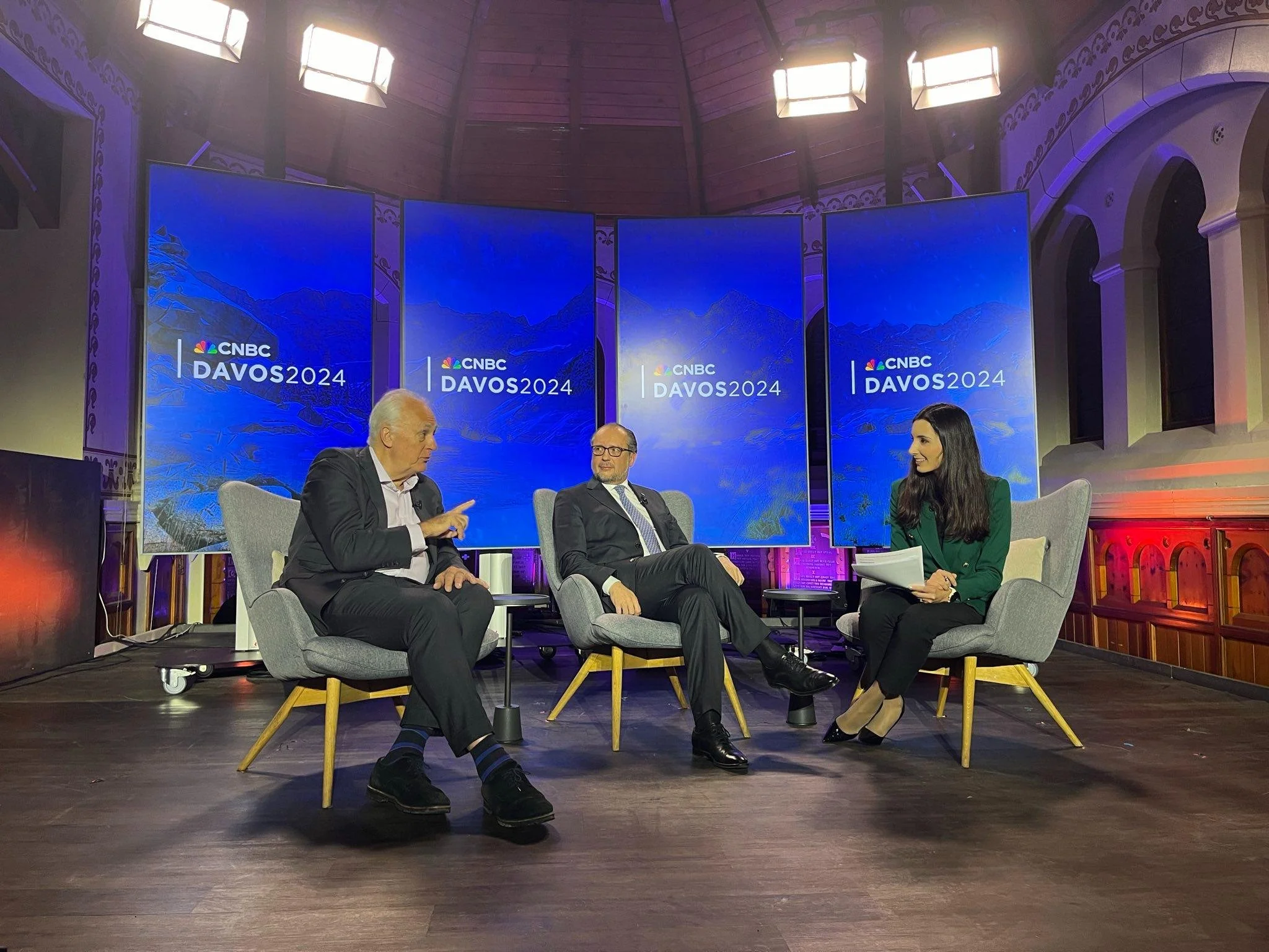 A panel discussion at the CNBC Davos 2024 event featuring three individuals seated on stage in front of three large blue screens with mountain graphics and the event logo. The room has ornate architectural details and wooden flooring.