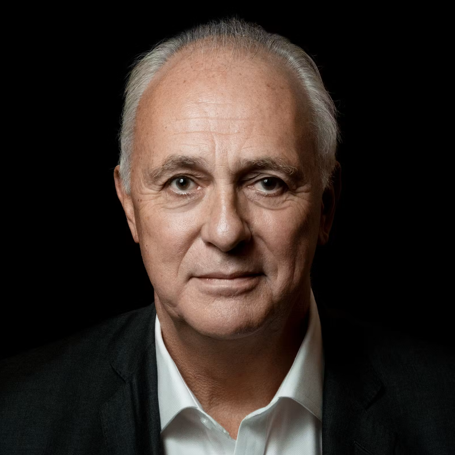 Close-up portrait of an older man with gray hair, wearing a white shirt and black suit jacket, against a black background.