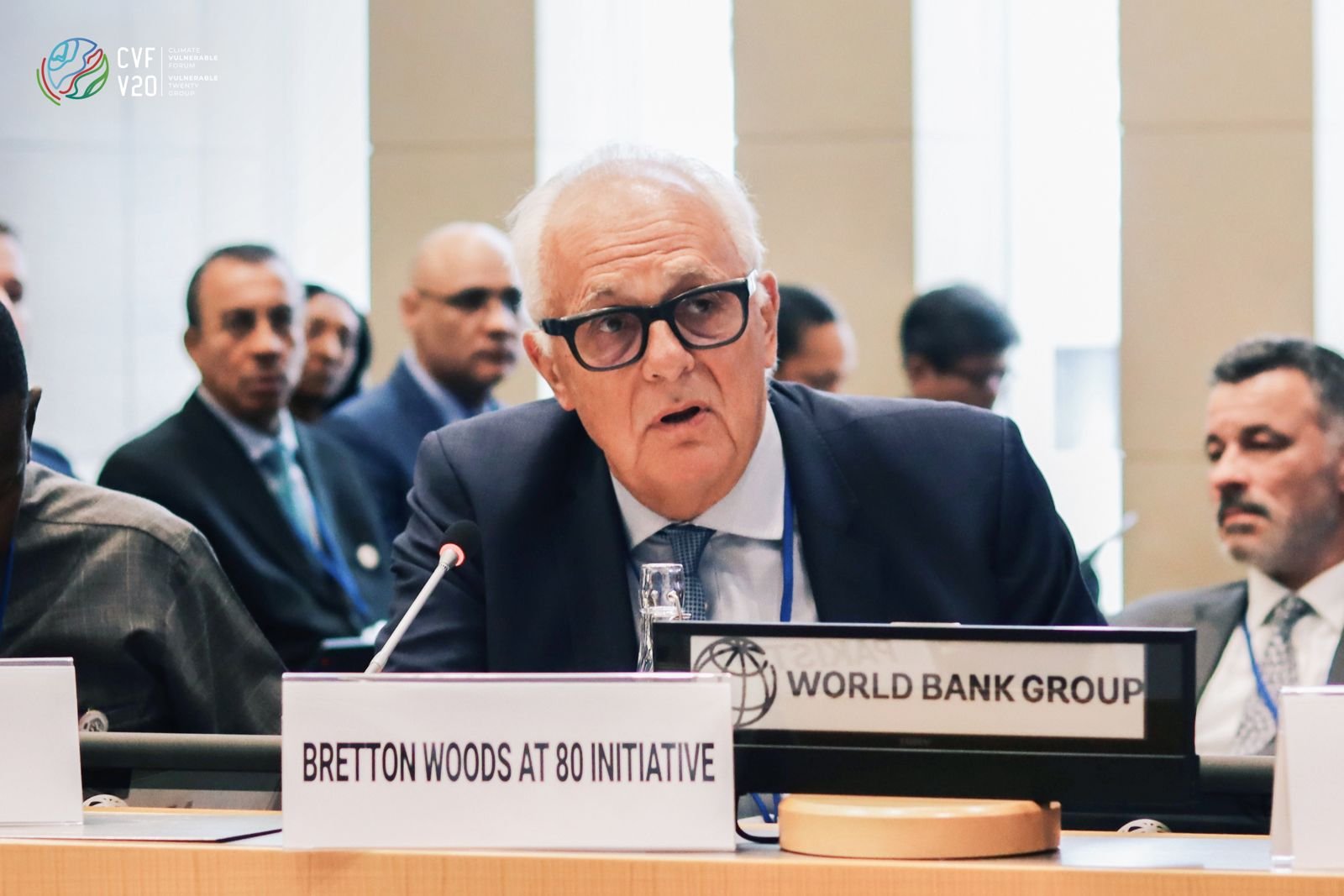 Bretton Woods conference meeting with a man speaking into a microphone. Nameplate reads 'Bretton Woods at 80 Initiative,' and a more prominent sign says 'World Bank Group.' Several individuals in business attire are seated around the table.