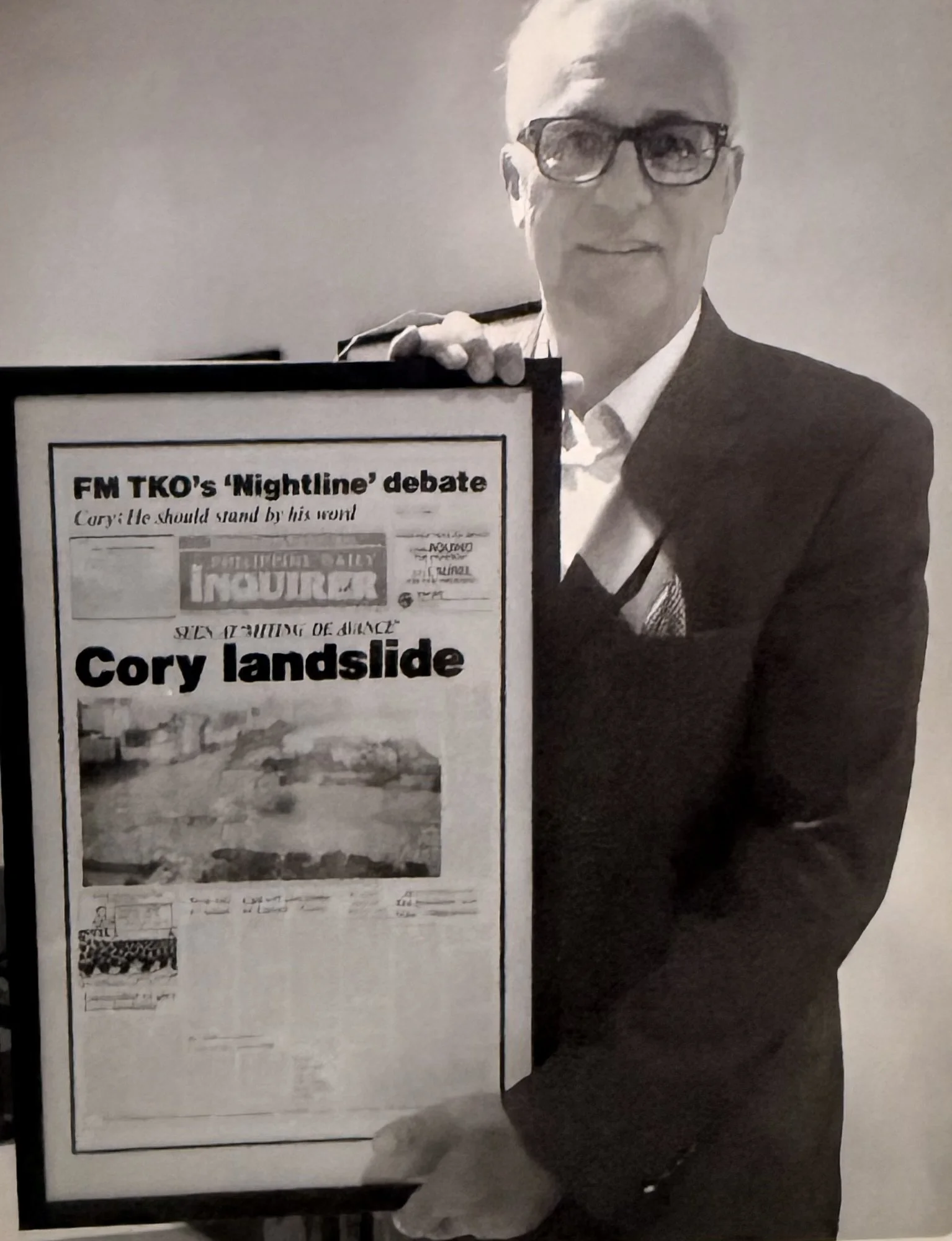 A woman with short hair, glasses, and a blazer holding a framed newspaper headline that reads, "Cory Landslide," at an indoor event.