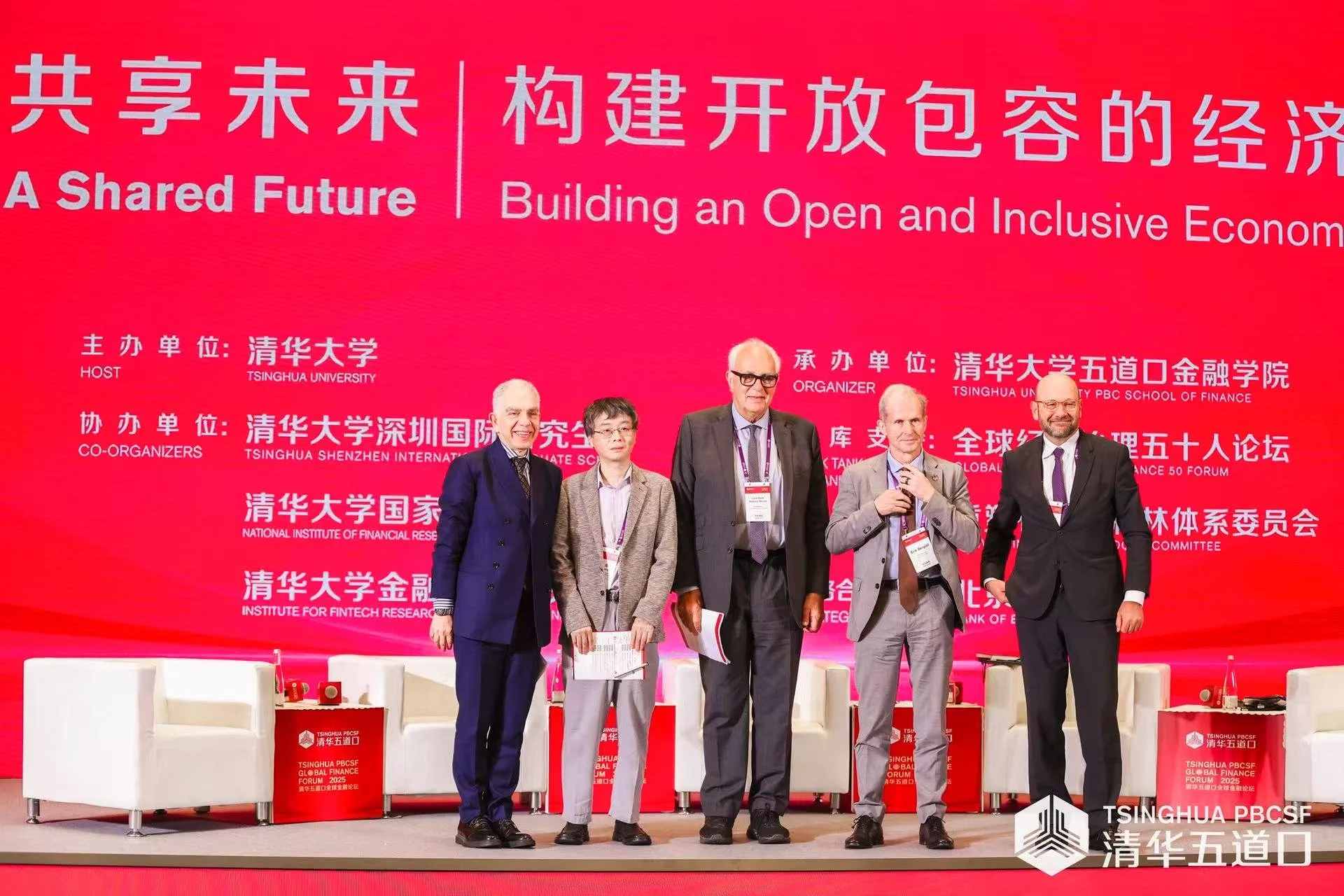 Group of five men in suits standing on stage at a conference, with a large red backdrop containing Chinese characters, English text about building an open and inclusive economy, and logos of Tsinghua University and other organizations.