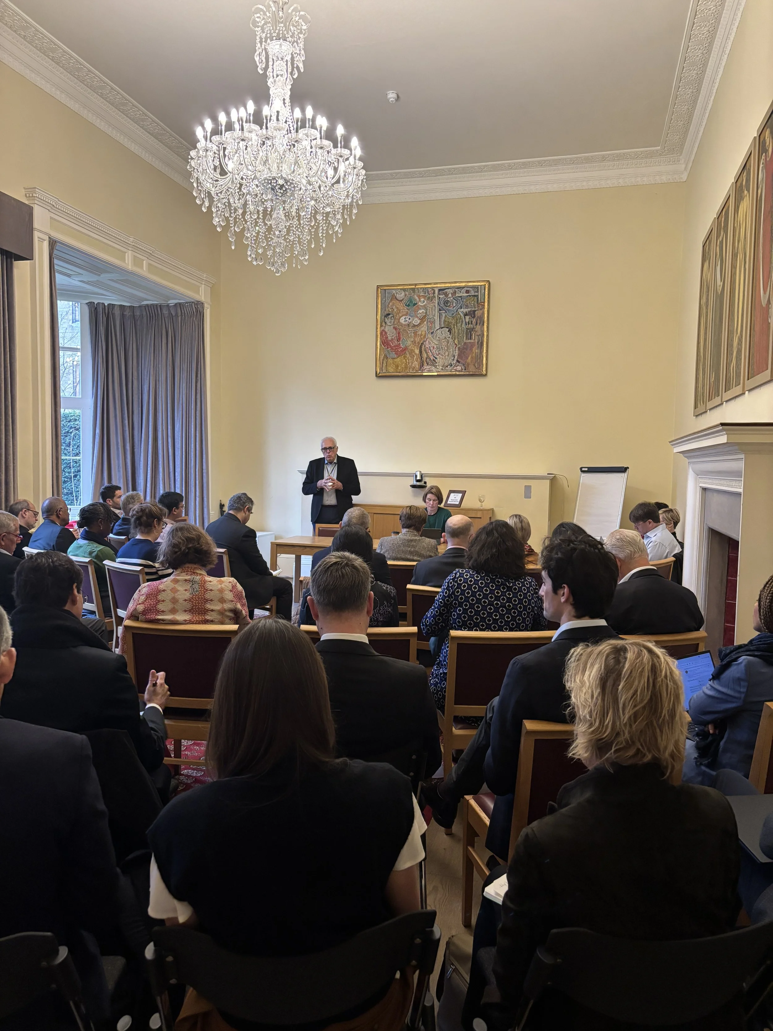 A group of diverse people gathered in a formal conference room with a high ceiling, ornate crown molding, and a large crystal chandelier. They are attending a presentation or lecture, with some taking notes or looking at a speaker at the front. The room has large windows with curtains and artwork hanging on the walls.