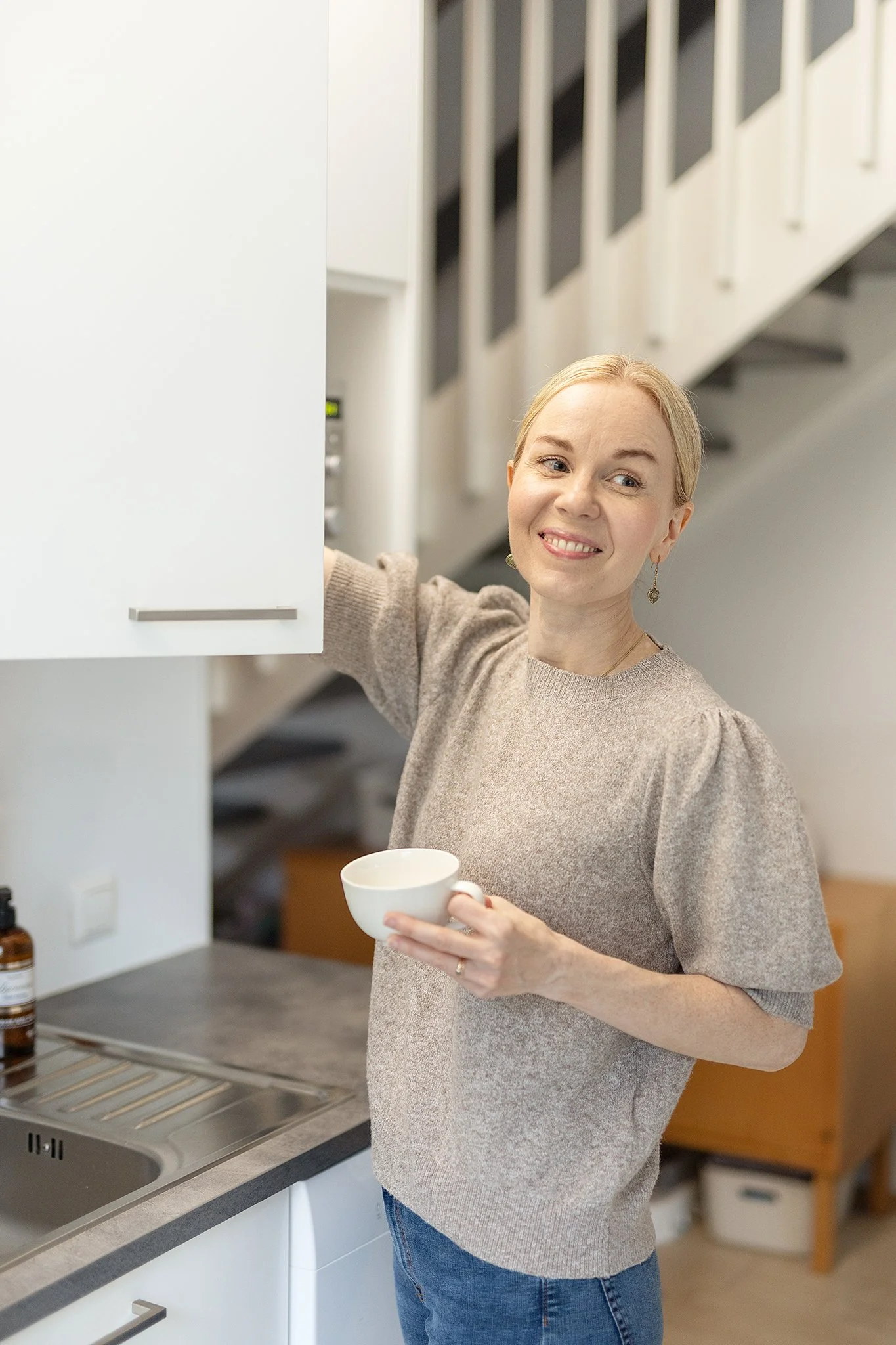 Nainen keittiössä, hänellä on vaaleat hiukset, ja hän on hymyilevä, hänellä on kuppi kädessään, keittiössä on astianpesukone ja ruokailuvälineitä näkyvissä.
