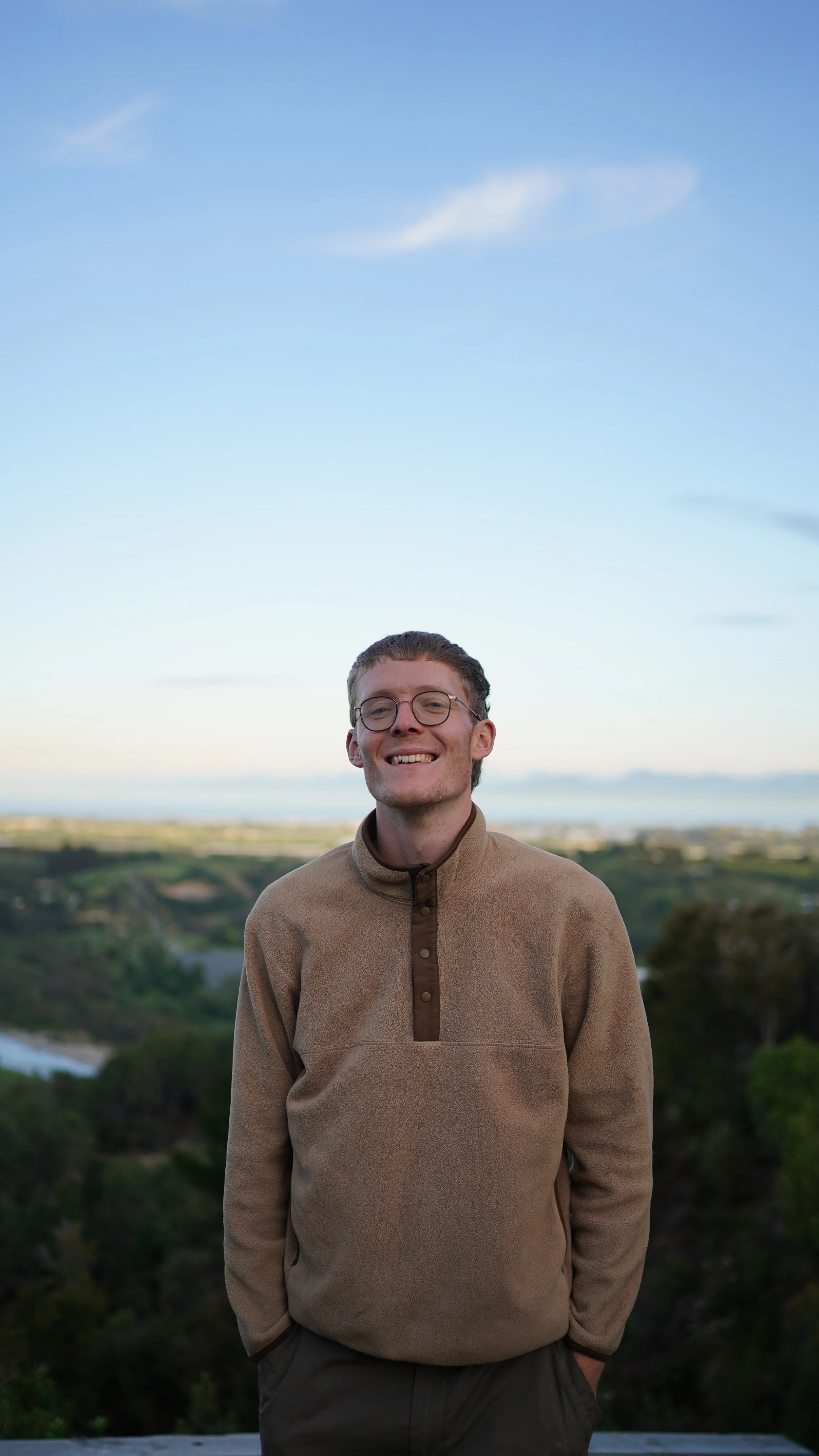 Young man with glasses smiling outdoors with hills and a body of water in the background during daytime.