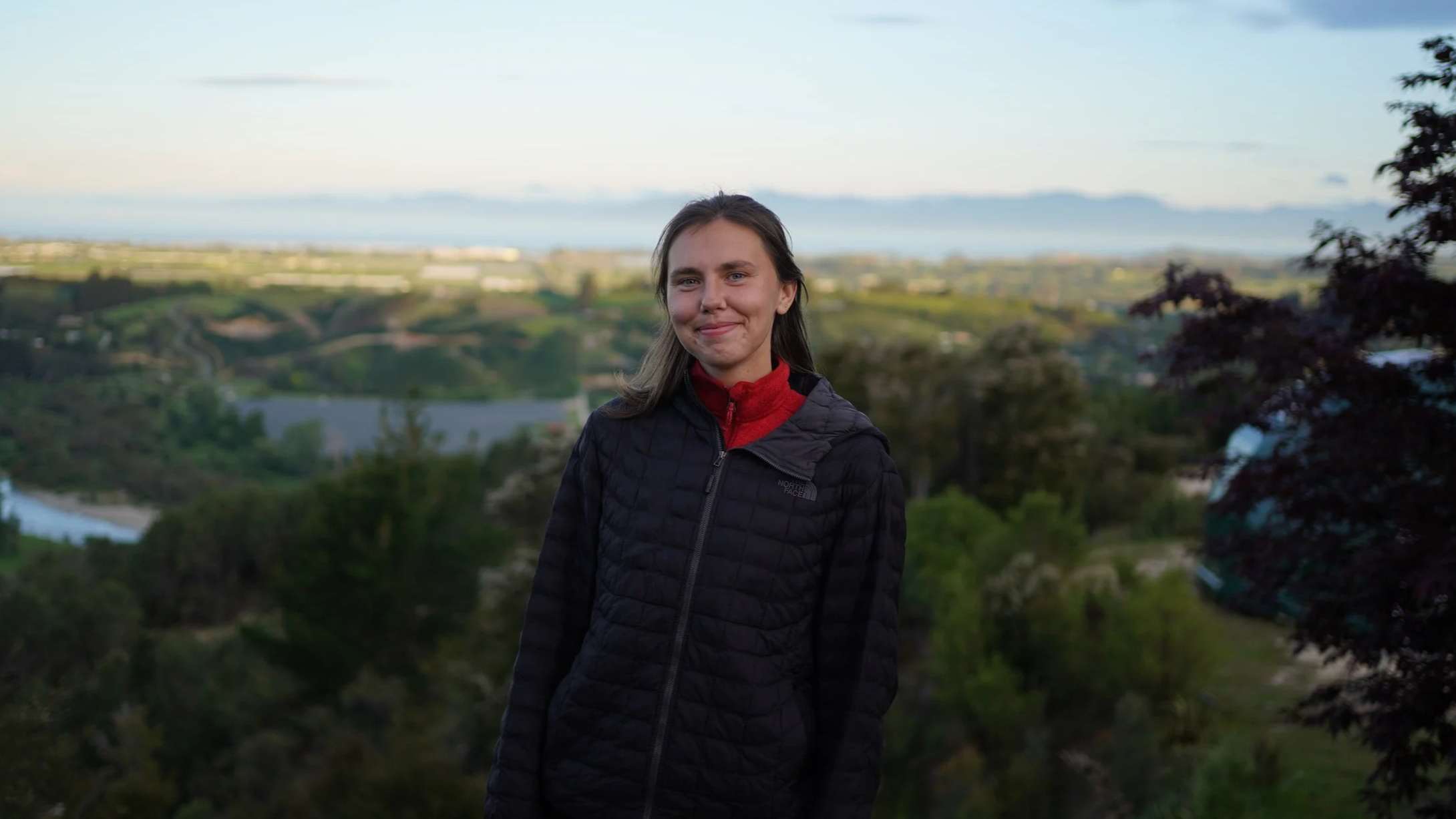 A young woman with long brown hair smiling outdoors, wearing a black jacket and red fleece, with a scenic landscape of hills, trees, and water in the background during the daytime.