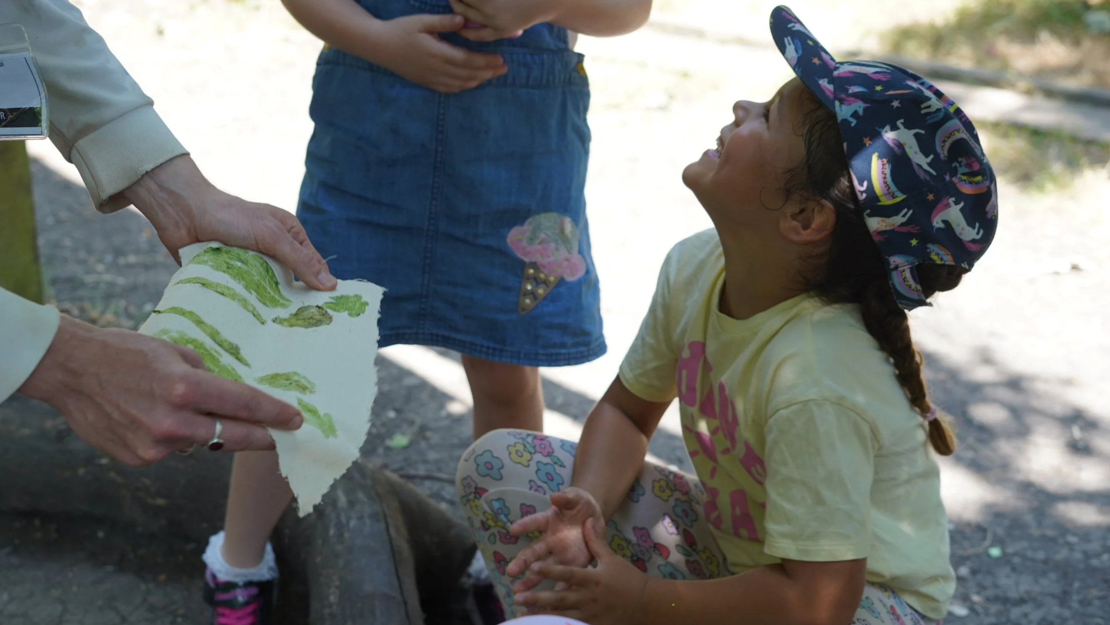 A young girl with a unicorn-themed hat sitting on a rock, smiling as she looks up at another person holding a piece of artwork. Two other children are standing nearby, one wearing patterned leggings and a yellow T-shirt.