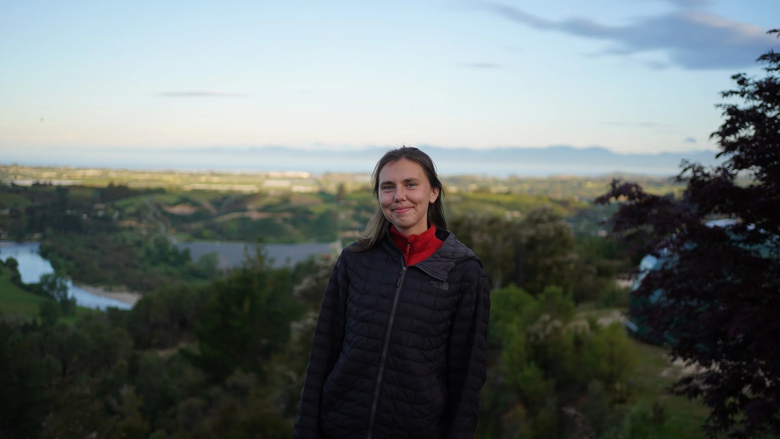A woman with brown hair smiling, wearing a black jacket and red shirt, standing outdoors with a scenic landscape of hills, trees, a river, and a cloudy sky in the background.