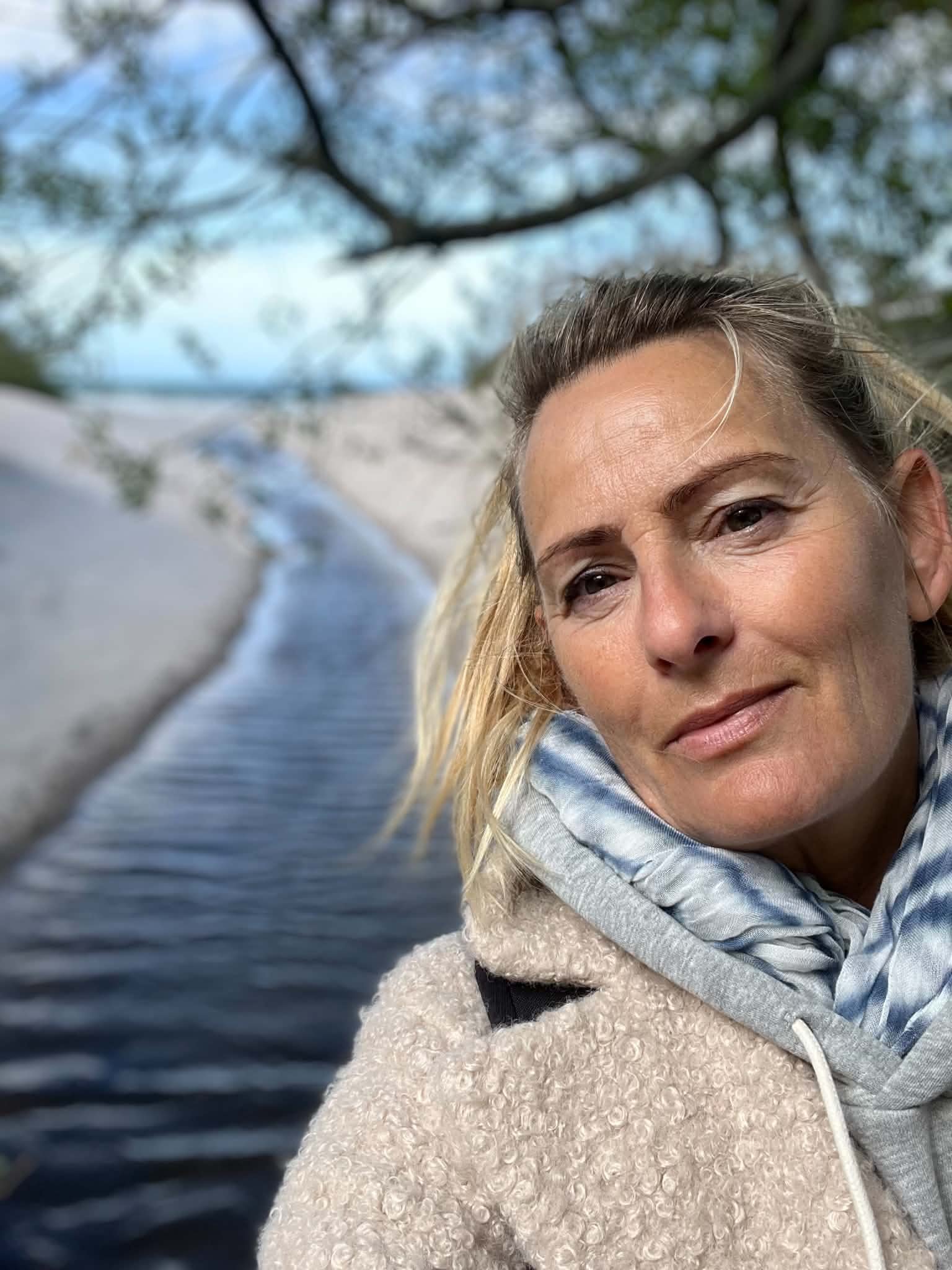 Nærbillede af en kvindes ansigt med blondt hår, iført en fluffy jakke og en lyseblå hoodie. Baggrund af en stor sø eller flod med træer og blå himmel.
