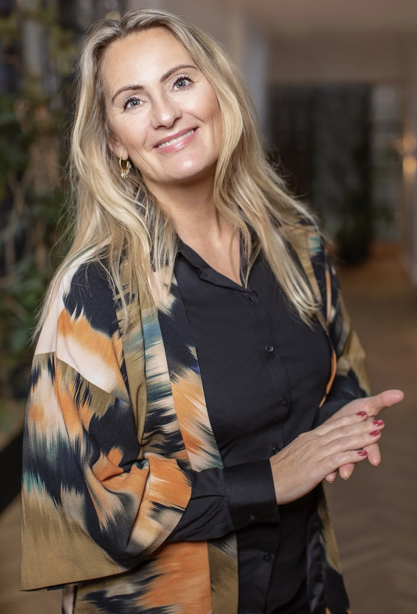 Kvindelig person med blondt, bølget hår, iført sort skjorte og en farverig kjole, smilende i indendørs miljø.