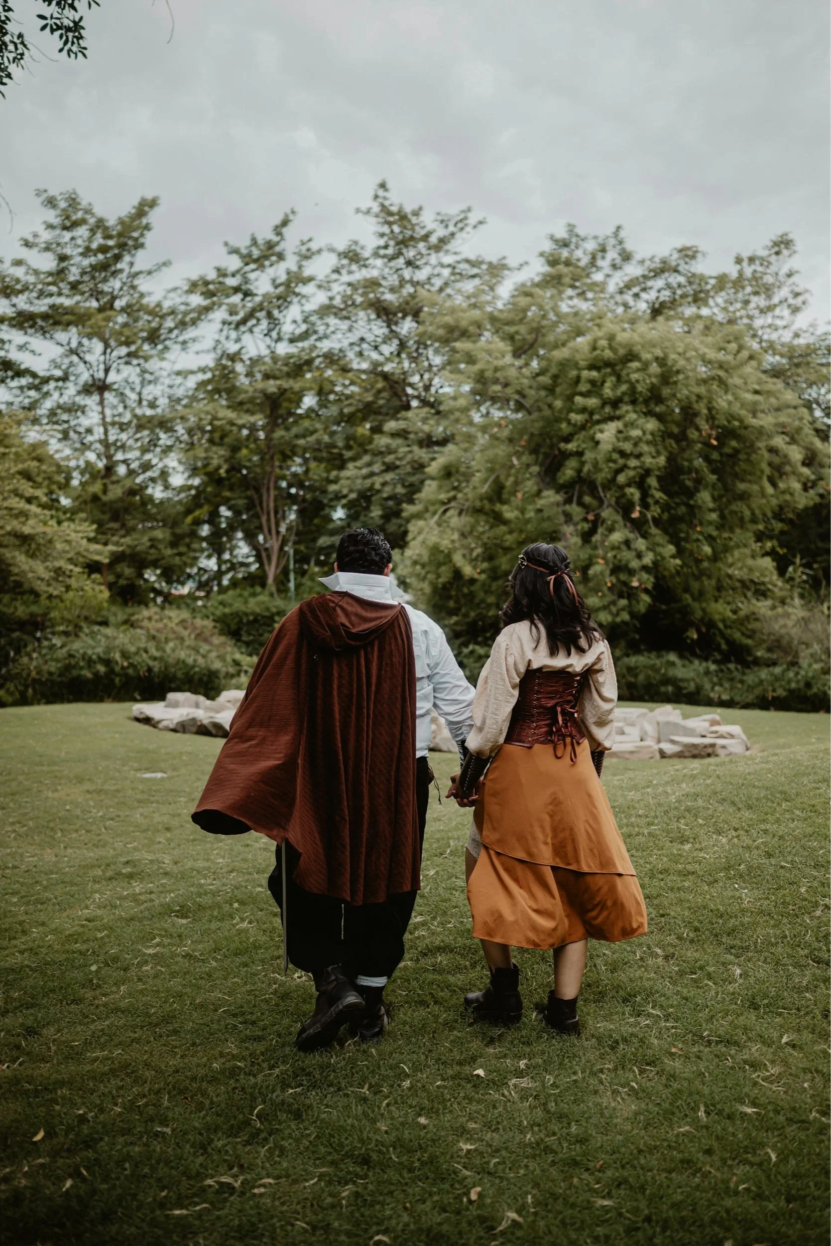Two people dressed in medieval or fantasy costumes walking on a grassy lawn with trees and cloudy sky in the background.
