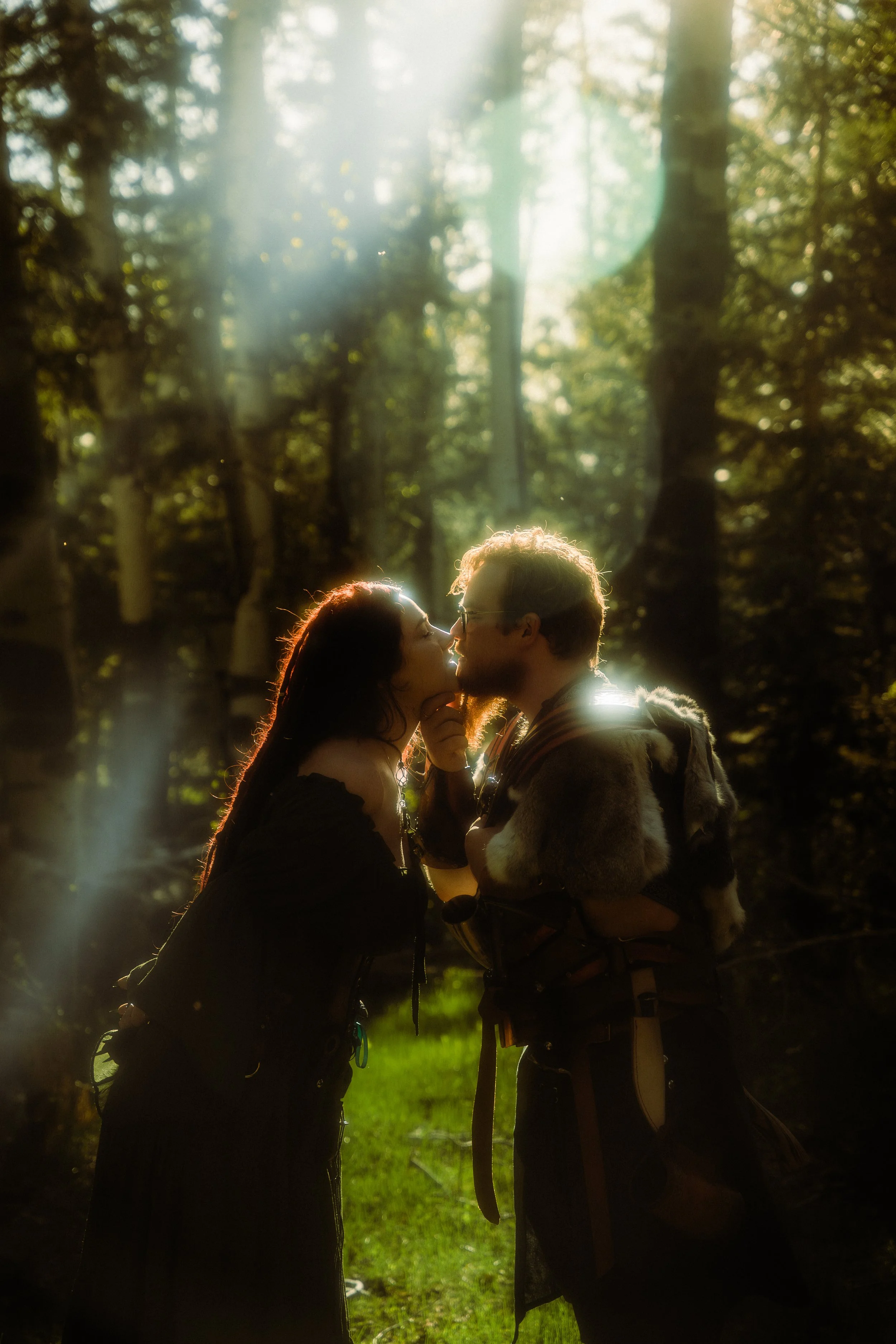 A couple is standing close together outdoors in a forest, touching foreheads and gazing into each other's eyes, with sunlight filtering through the trees in the background.