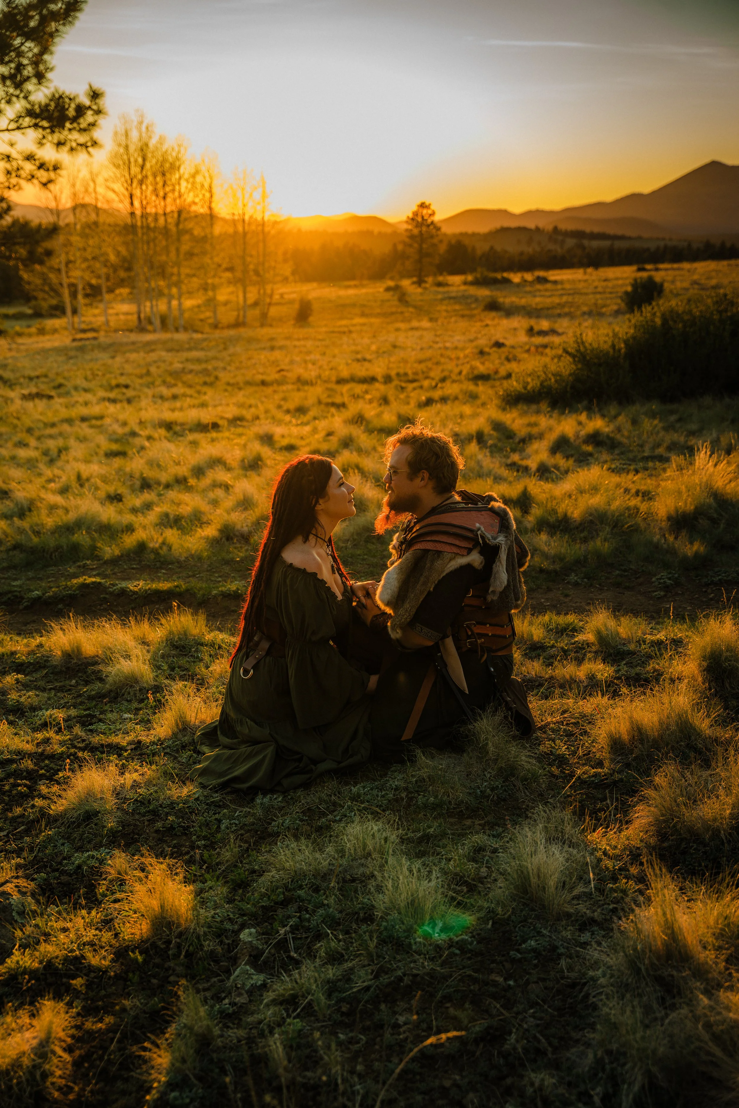 A man and woman kneeling in a grassy field during sunset, face to face, with mountains and trees in the background.