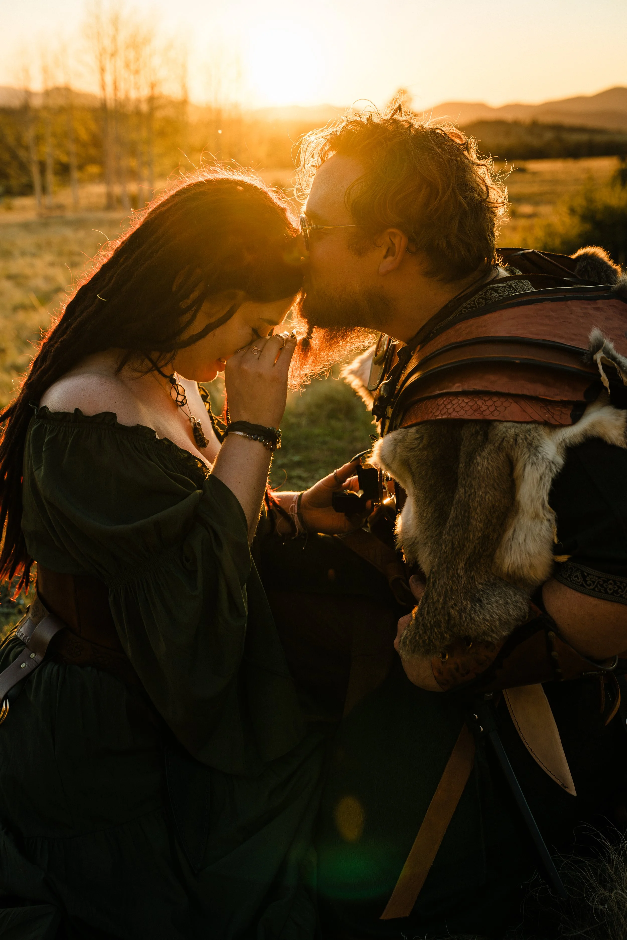 A woman and a man sharing a kiss outdoors during sunset. The woman has long braided hair and wears black clothing with jewelry. The man has curly hair, glasses, and is dressed in medieval or fantasy-style armor with fur accents, holding a sword. The background shows a rural landscape with leafless trees and mountains.