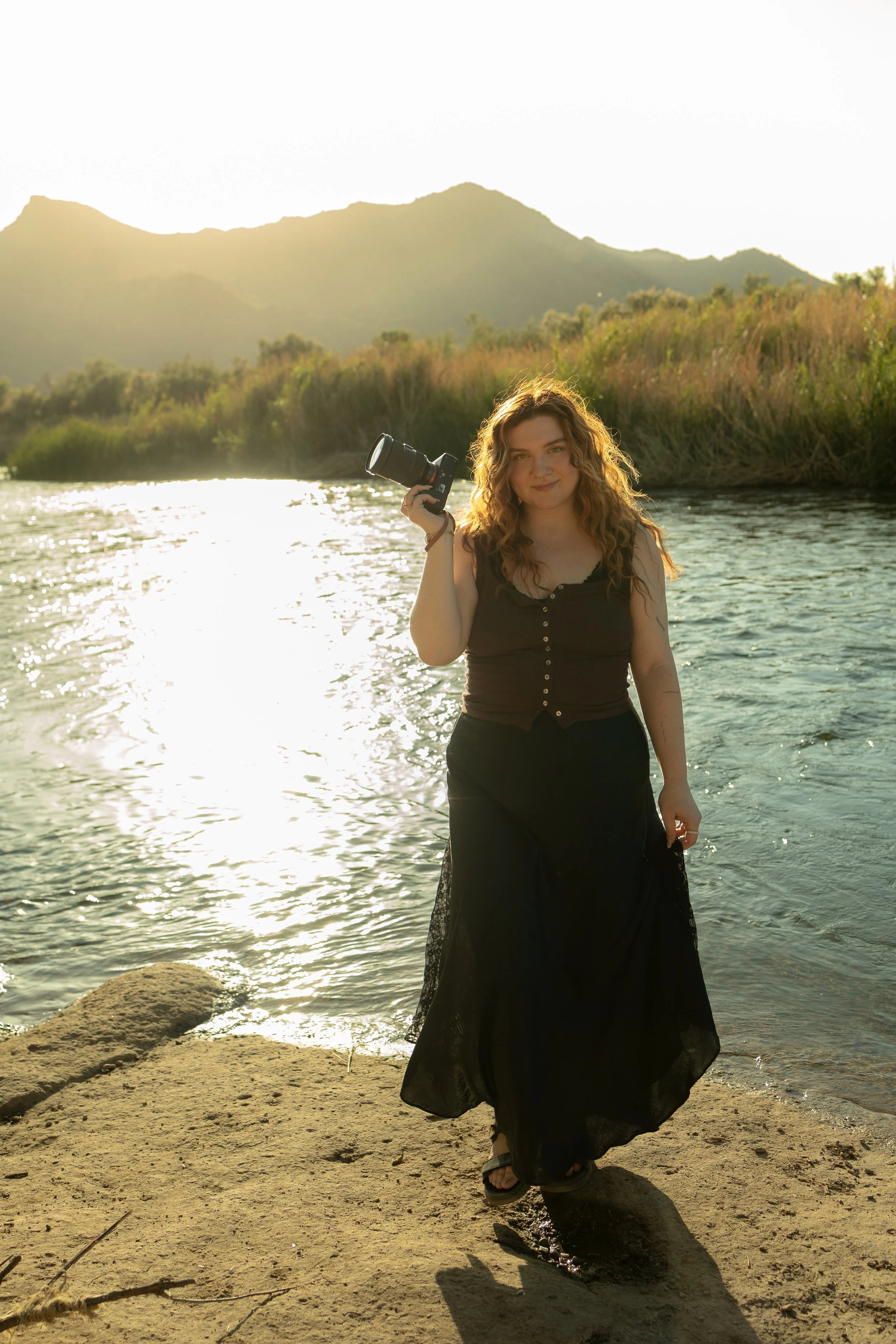 A woman with curly hair standing on a rock by a river, holding a camera, with a mountain landscape in the background during sunset.