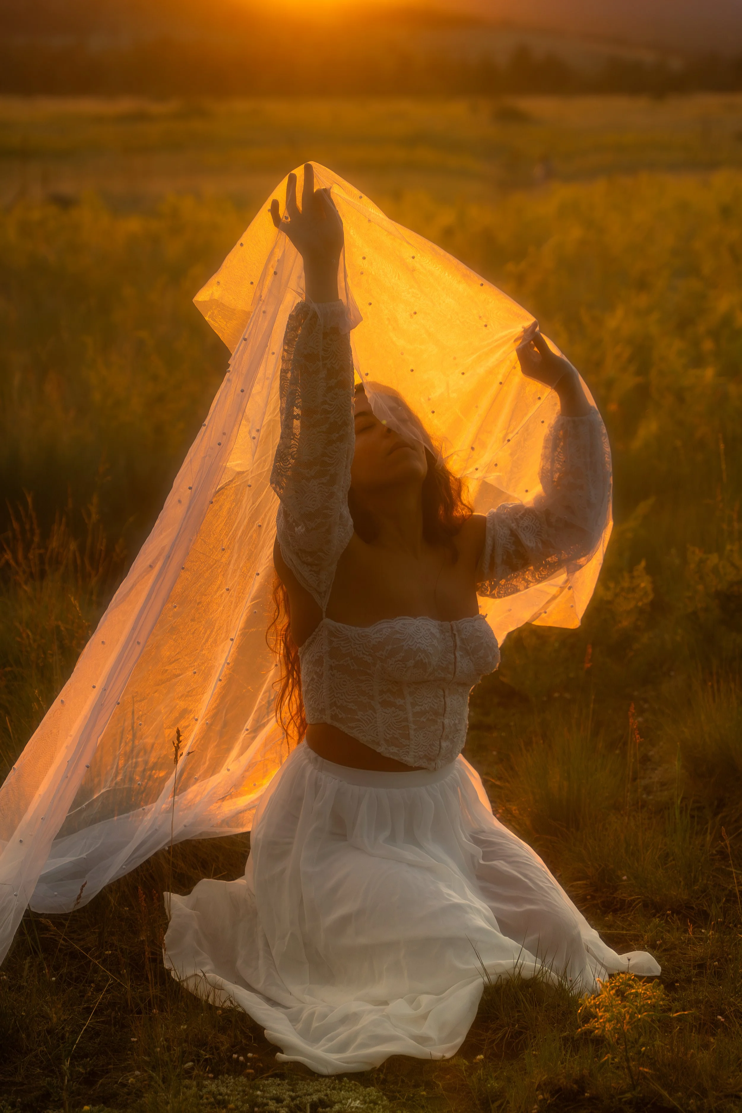 A woman kneeling on the grass holding a sheer orange fabric up to the setting sun in a field at sunset, wearing a white lace crop top and a long white skirt.
