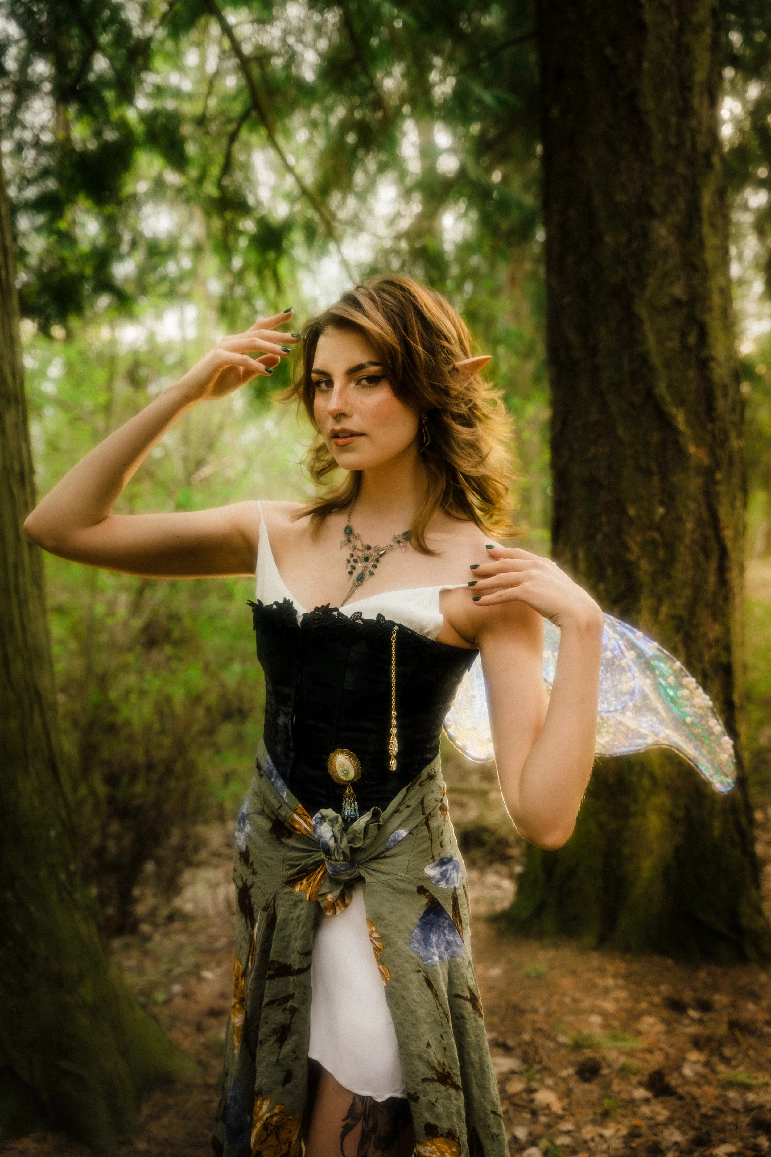 A woman dressed as a fairy with butterfly wings, elf ears, and a fantasy-themed necklace standing in a forest.
