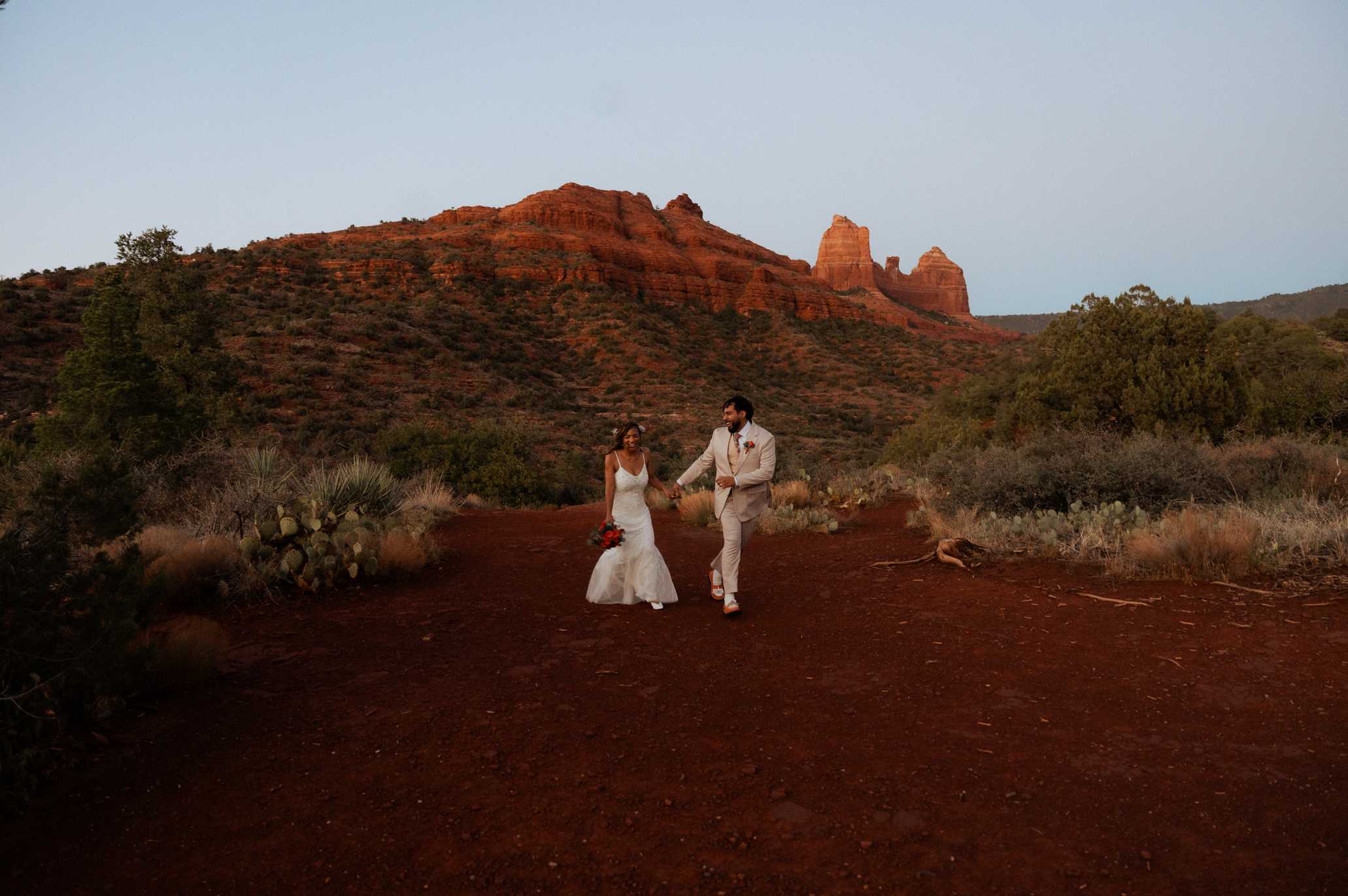 A couple dressed in wedding attire walking hand-in-hand on a dirt trail in a desert landscape with red rock formations and sparse green vegetation.