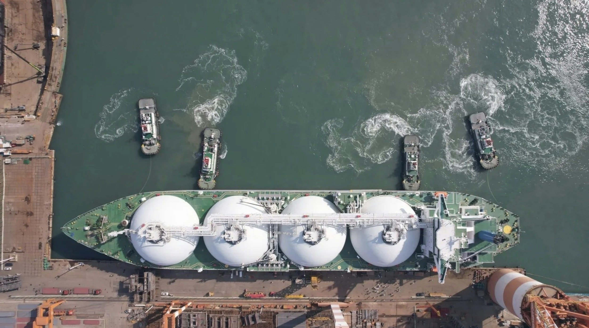 Aerial view of a large ship, possibly a cargo or industrial vessel, docked at a port with ships and water surrounding it.