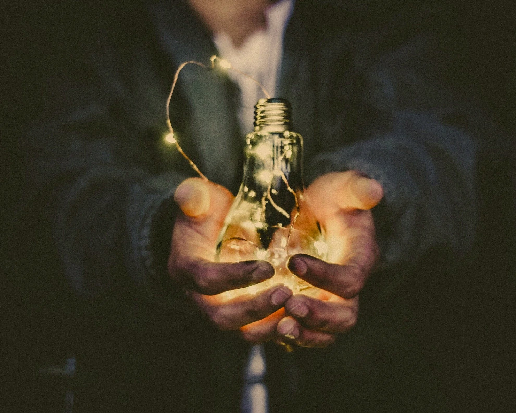 Person holding a clear lightbulb with string lights inside of it in a dark setting.