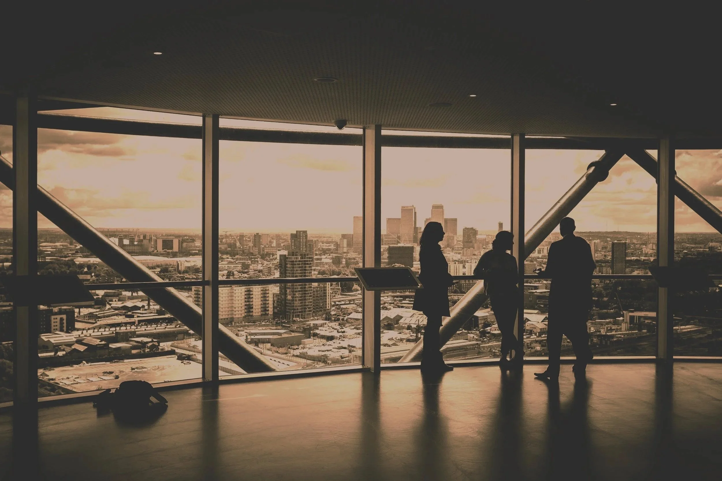 Silhouettes of three people standing and talking inside a high-rise building with large windows overlooking a city skyline at sunset.