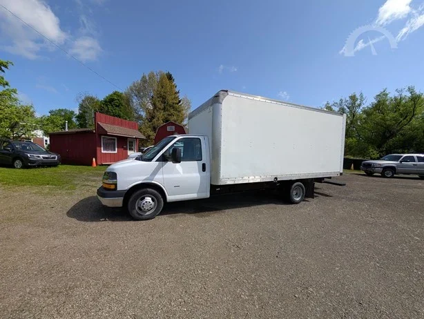White box truck parked on a gravel lot with trees and small red building in the background