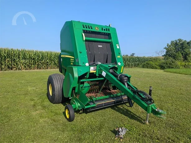 Green John Deere hay baler in a field with a small animal in front and a backdrop of crops and trees.