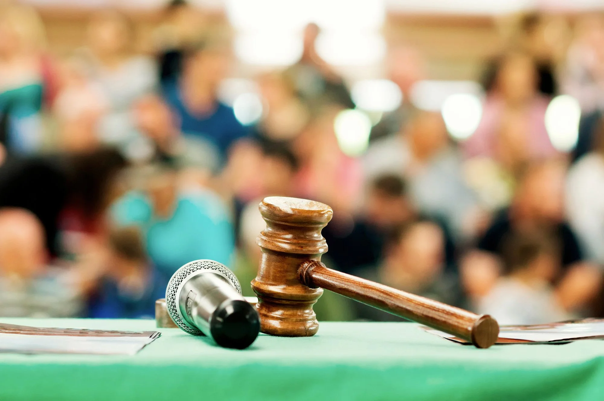 A judge's gavel, a microphone, and a document on a green table in a courtroom with people in the background.