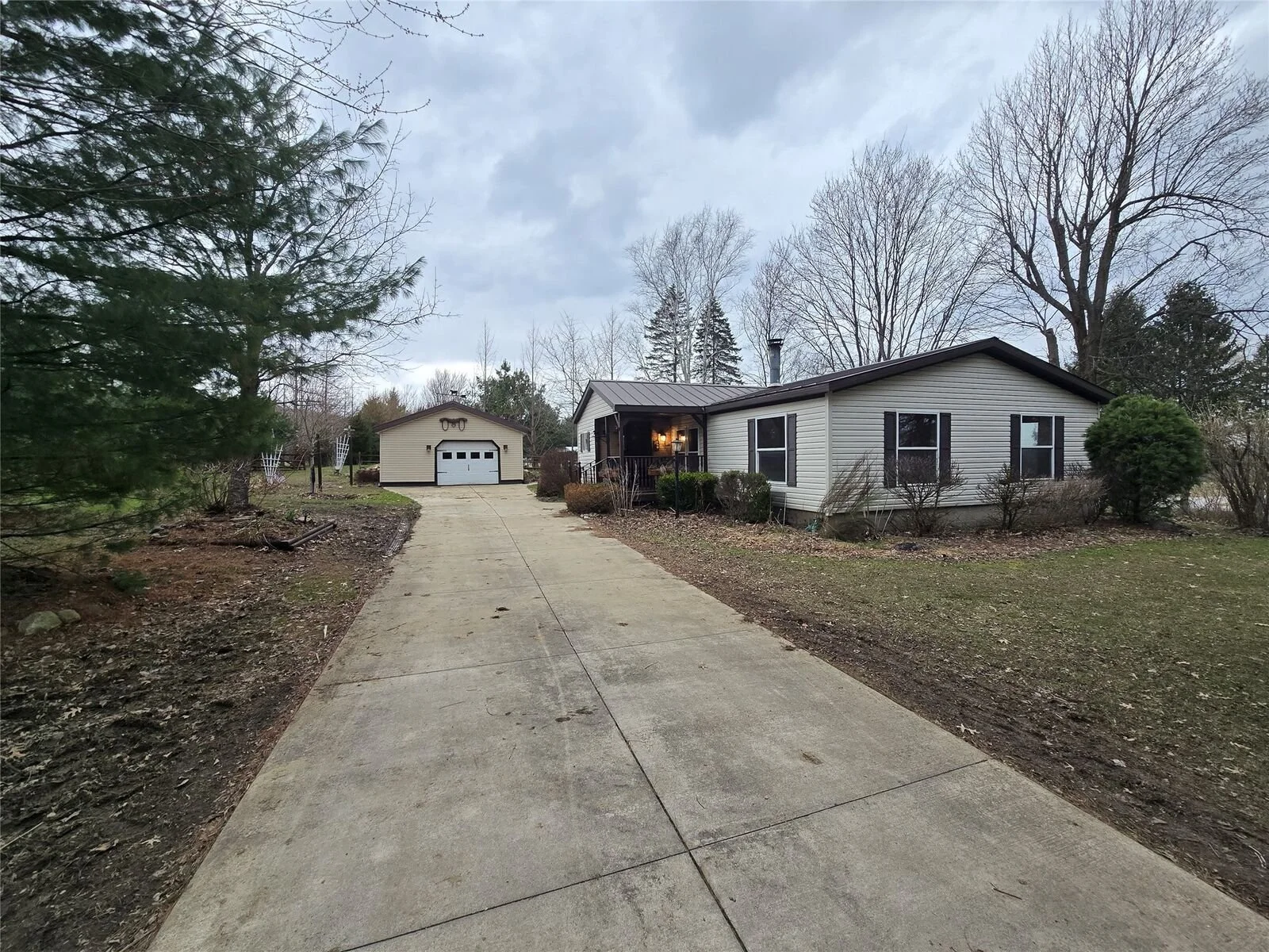 A single-story house with a small front porch and a detached garage at the end of a concrete driveway. There are leafless trees and bushes surrounding the house, with a cloudy sky overhead.