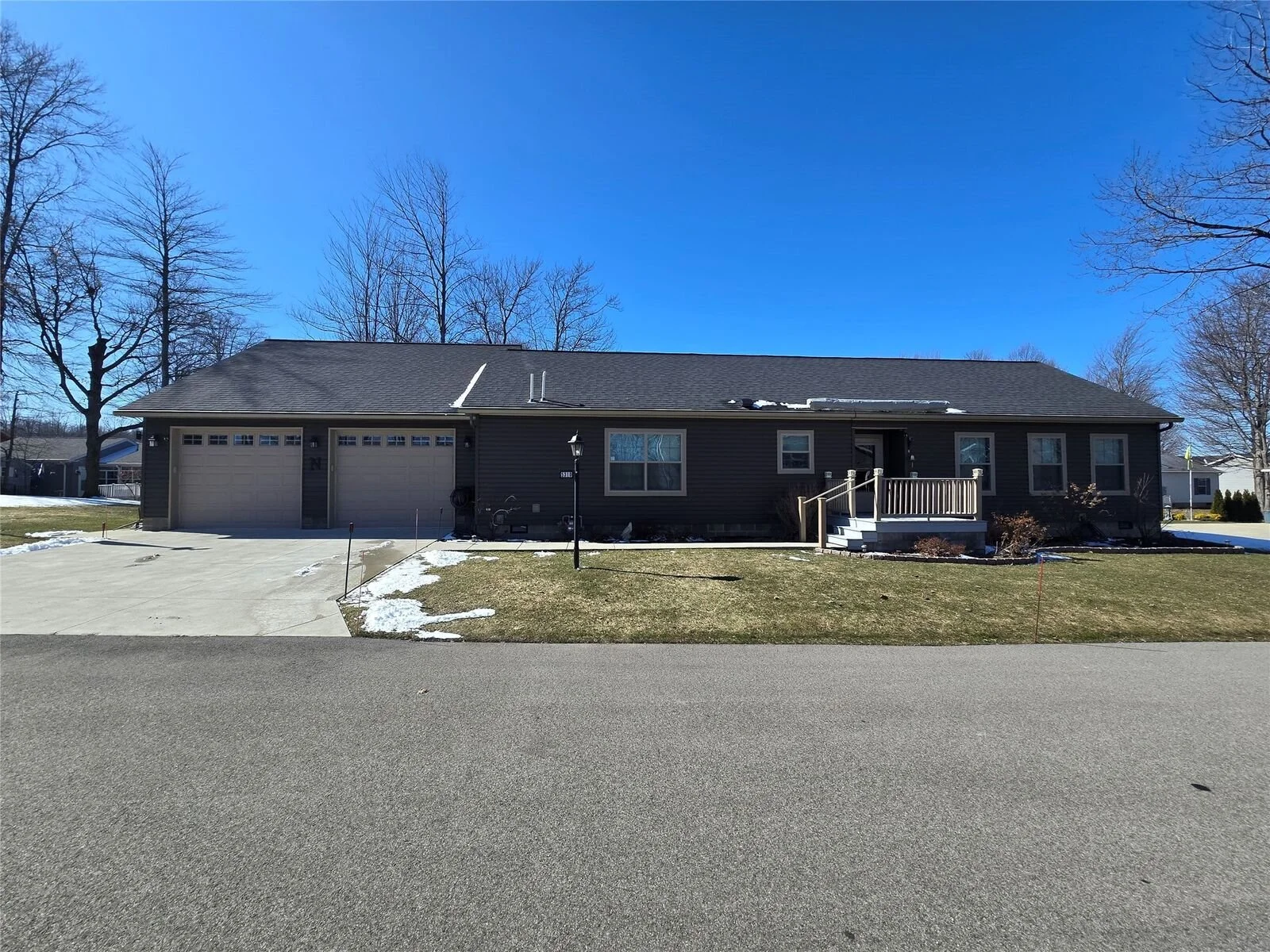 A single-story house with gray siding, a two-car garage, a small elevated front porch with white railings, a gray roof with some snow, on a partly snowy lawn, under a clear blue sky, with leafless trees in the background.