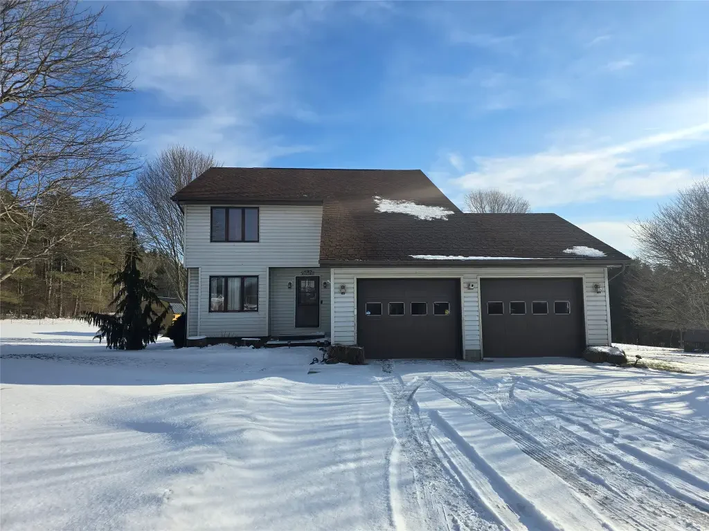 A two-story house with white siding and a brown roof, with snow on the ground and on parts of the roof, under a blue sky with some clouds.