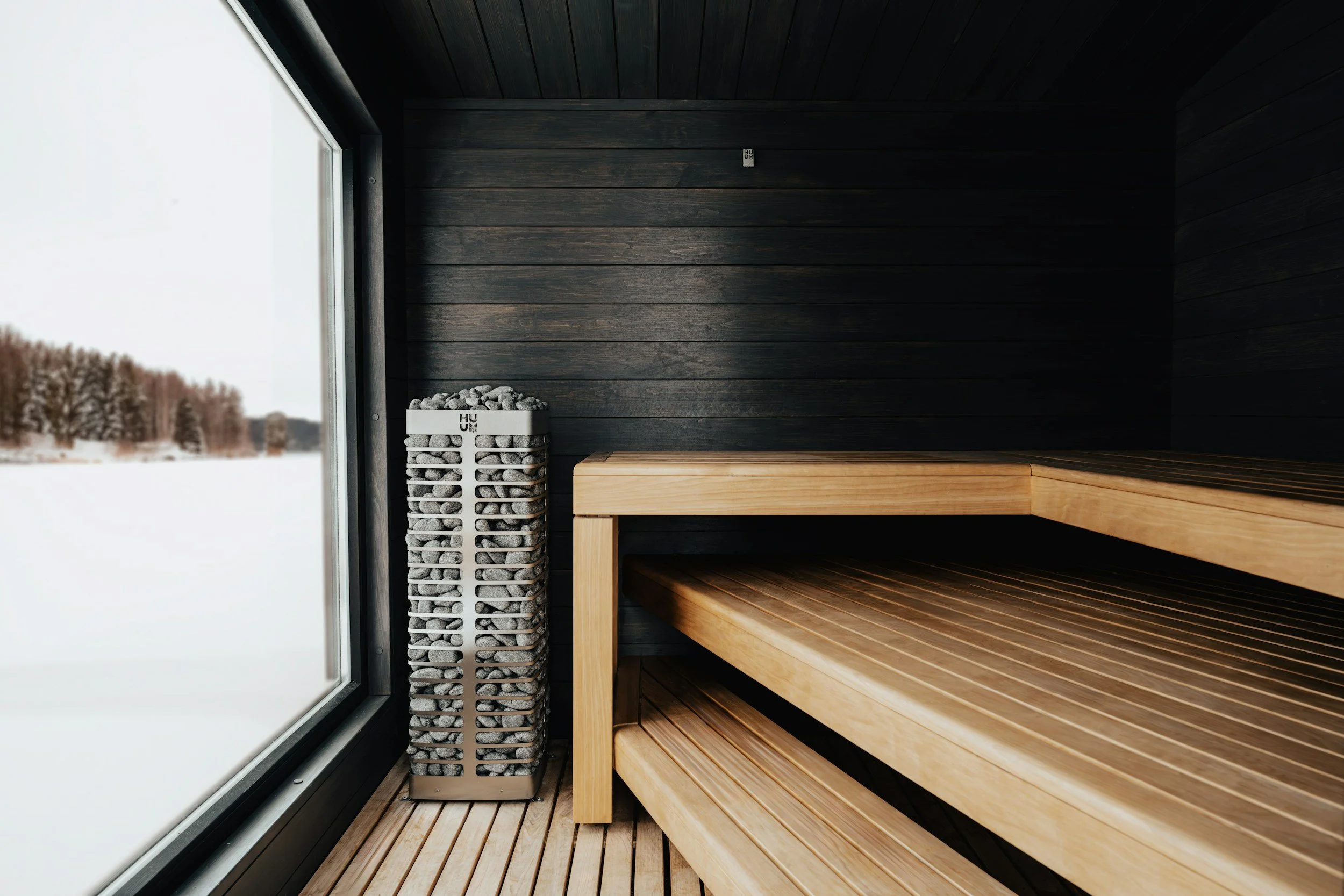 Interior of a modern sauna with wooden benches, black wood-paneled walls, a basket of stones, and a large window showing a snowy landscape outside.