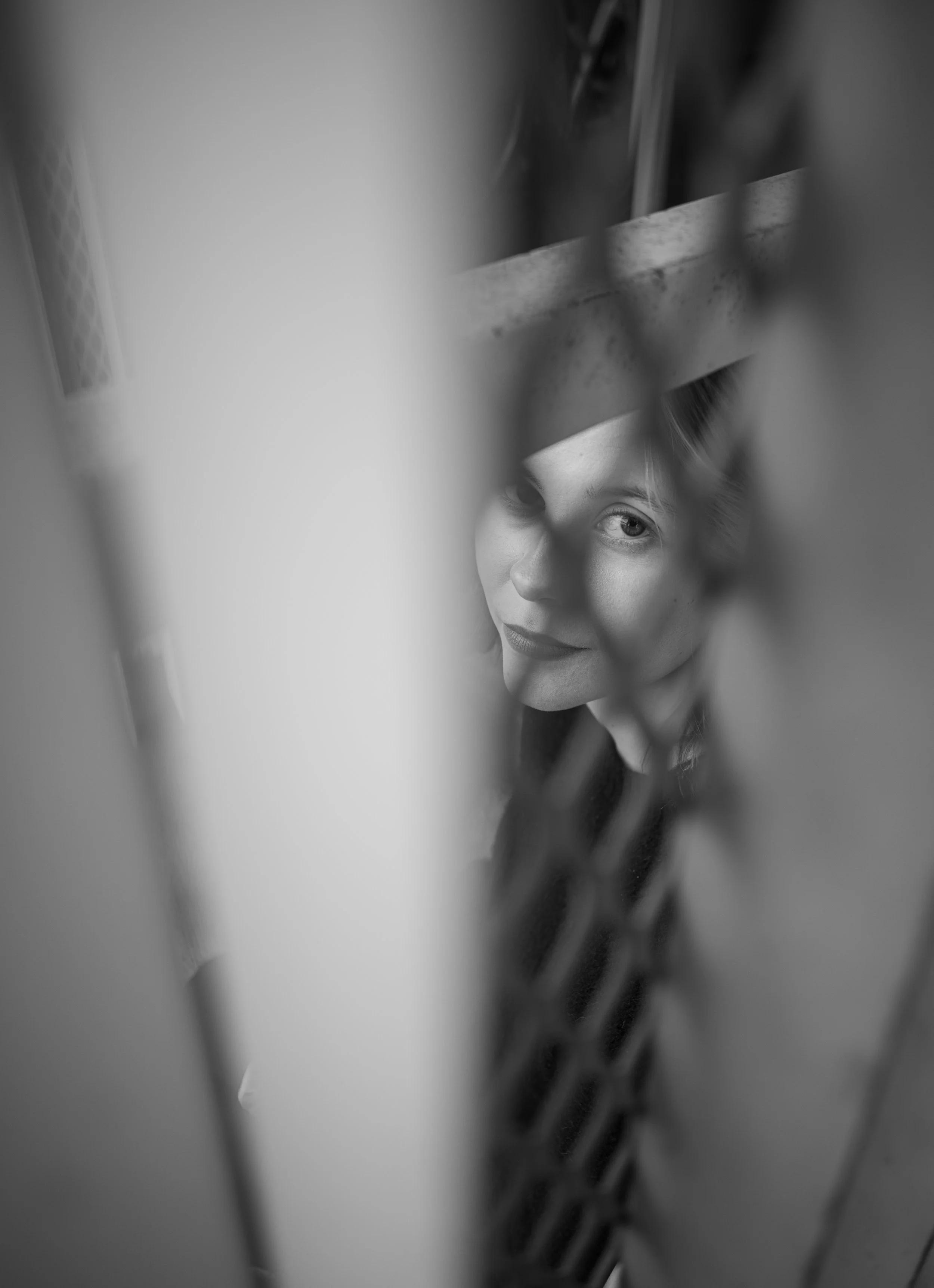 Black and white photo of a young woman peering through the bars of a metallic fence.