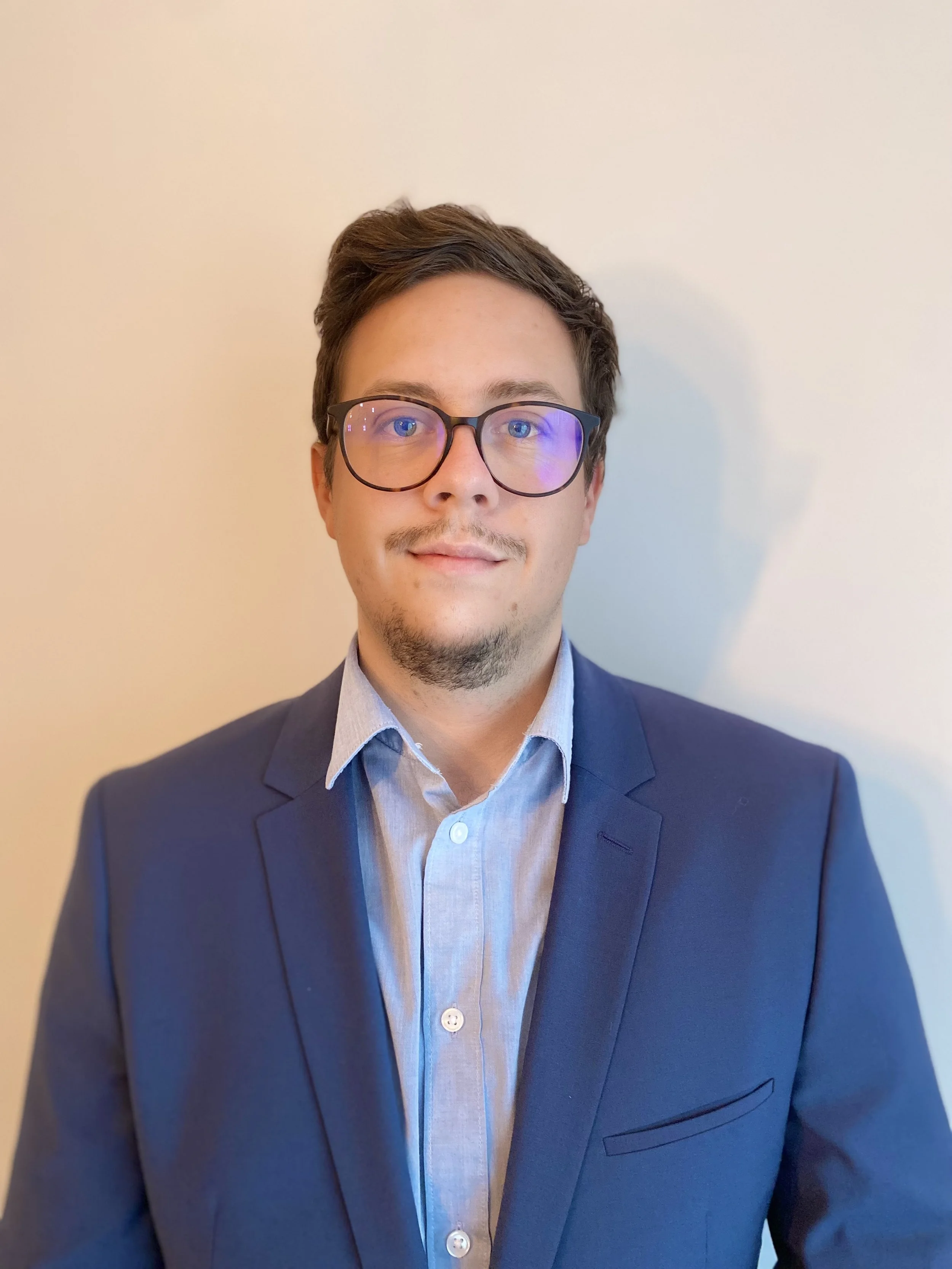 Mann mit hellbraunem Haar, brauner Brille und blauem Anzug lächelt in die Kamera.