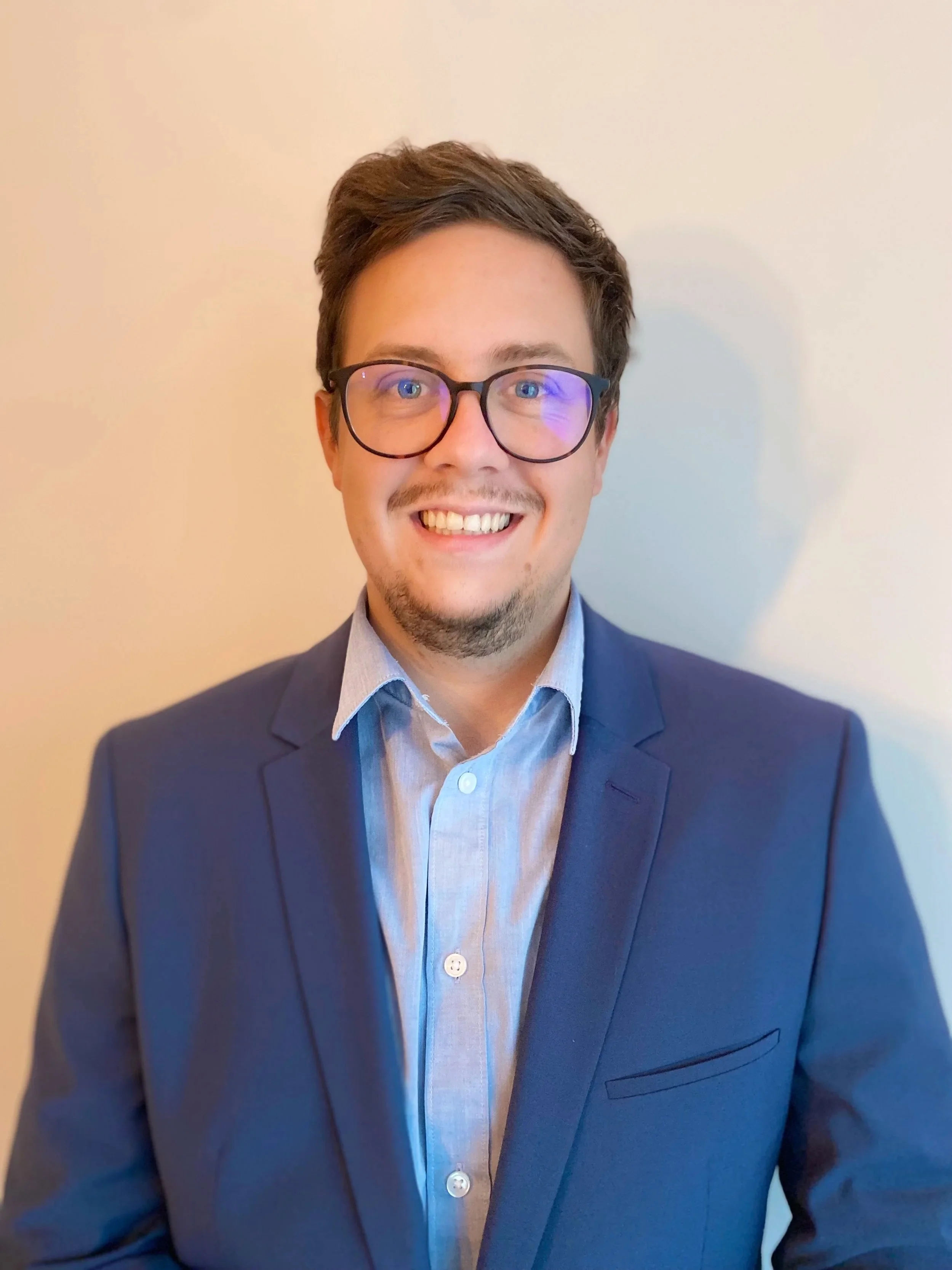 Mann mit hellbraunem Haar, Brille und blauem Anzug lächelt in die Kamera.