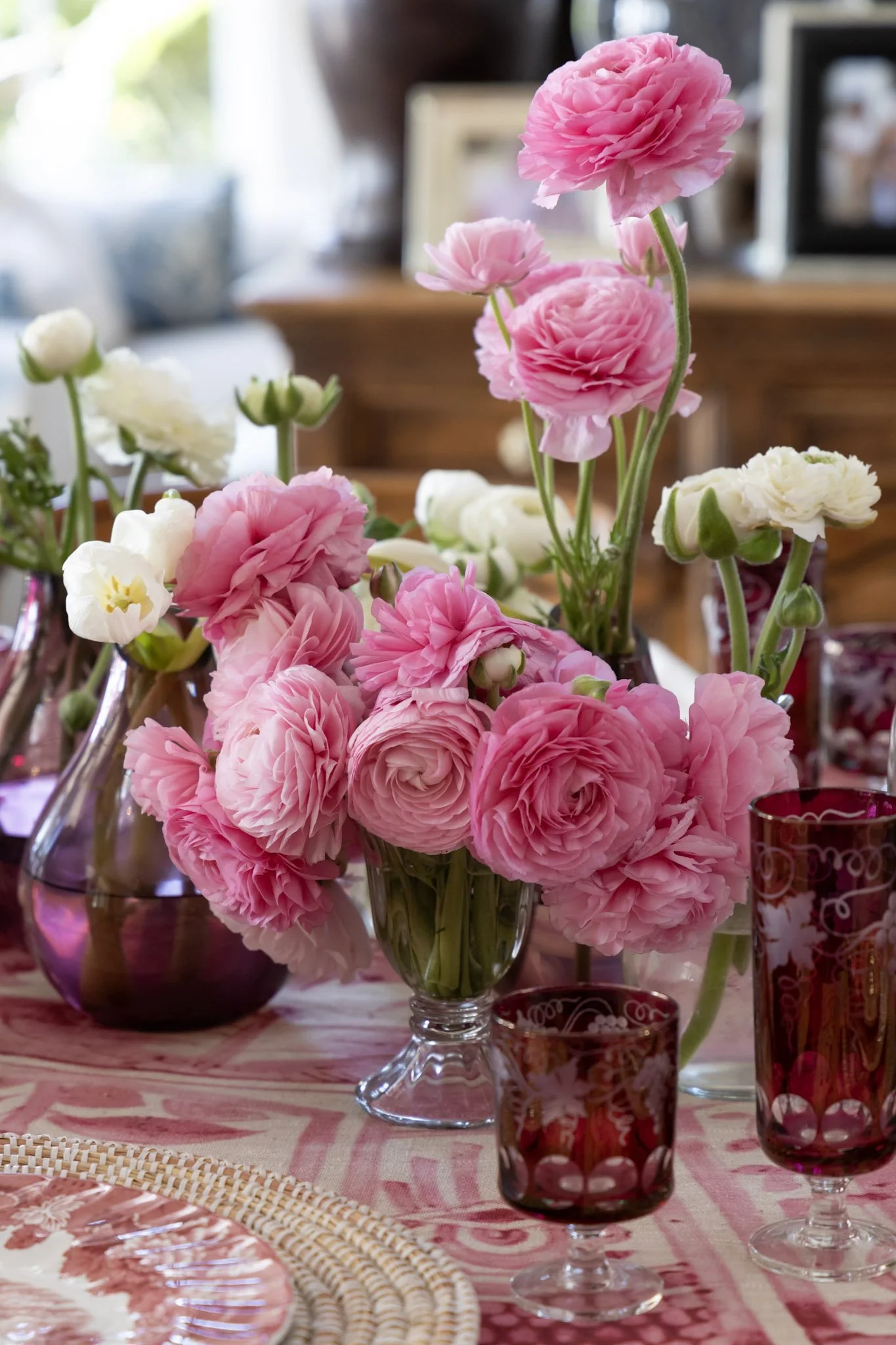 A glass vase filled with pink and white ranunculus flowers arranged on a table with pink and red decor, including glassware and a tablecloth with a pink pattern.