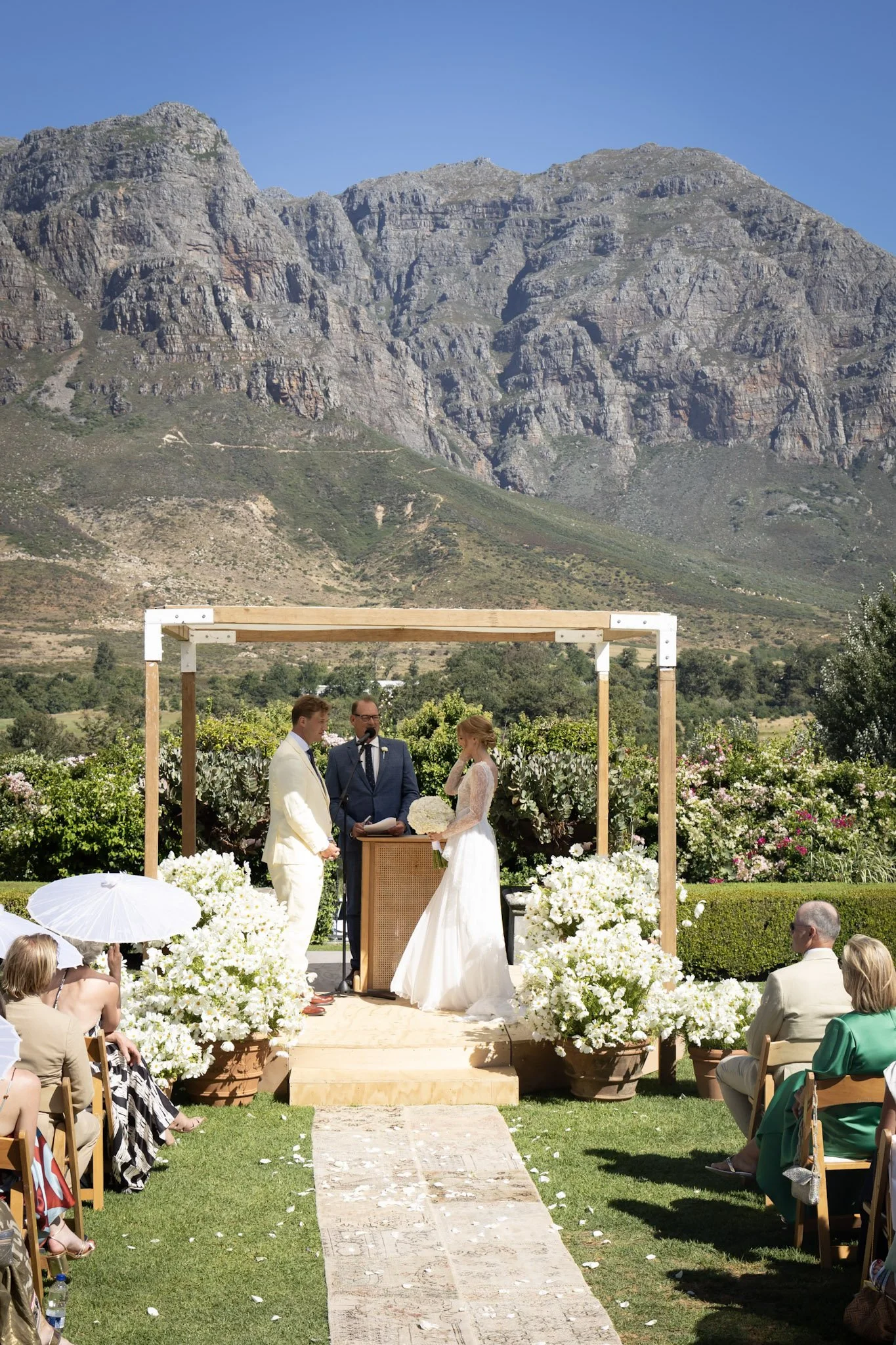 A wedding ceremony outdoors with a mountainous backdrop, an officiant, a groom in a white suit, a bride in a white dress, and guests seated on both sides of a grassy aisle decorated with white flowers and petals.