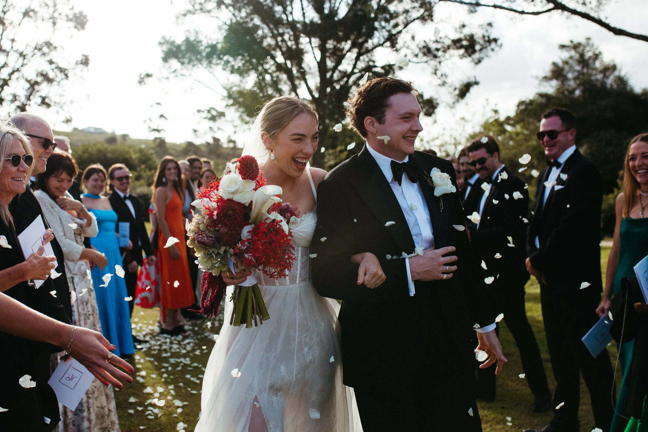 A bride and groom smiling and walking down the aisle during a wedding, surrounded by guests, with flower petals falling.