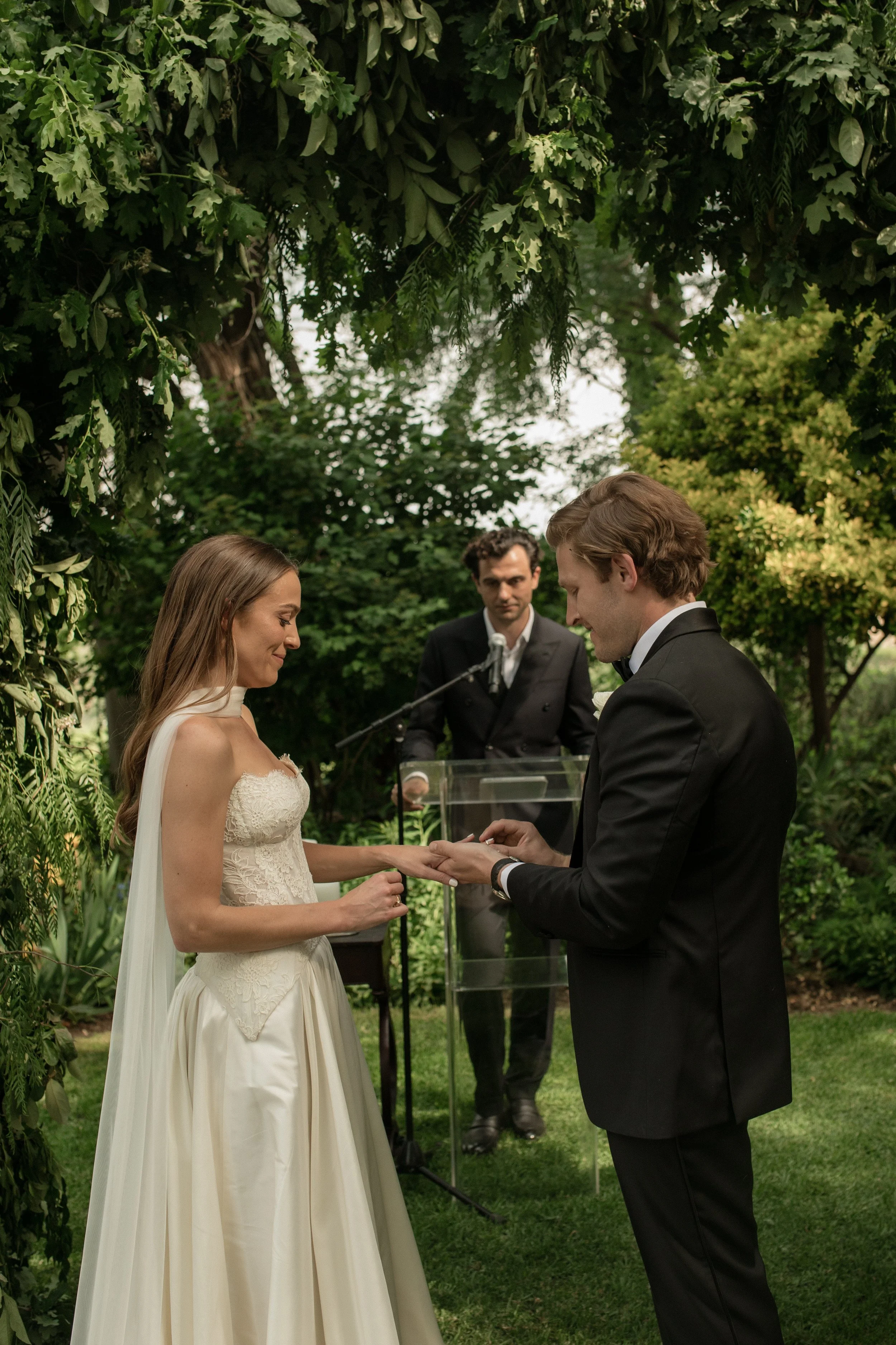A bride and groom exchanging rings during an outdoor wedding ceremony, with a minister officiating, under a leafy green arch.