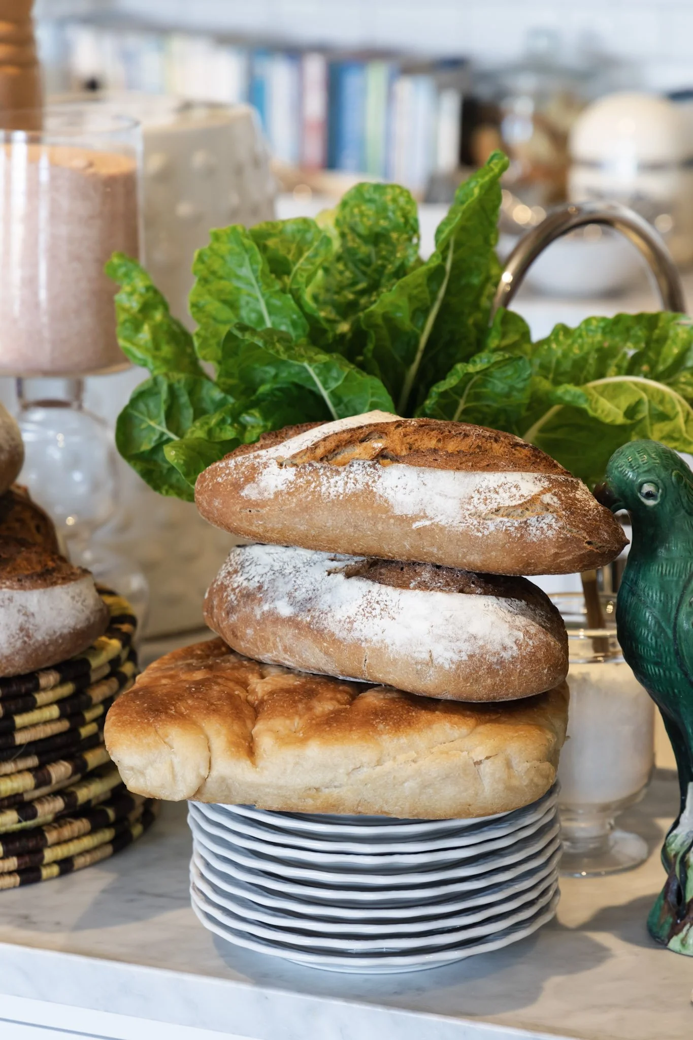 Stacked bread rolls and a large pastry on a striped plate, with leafy greens and other baked goods in the background.