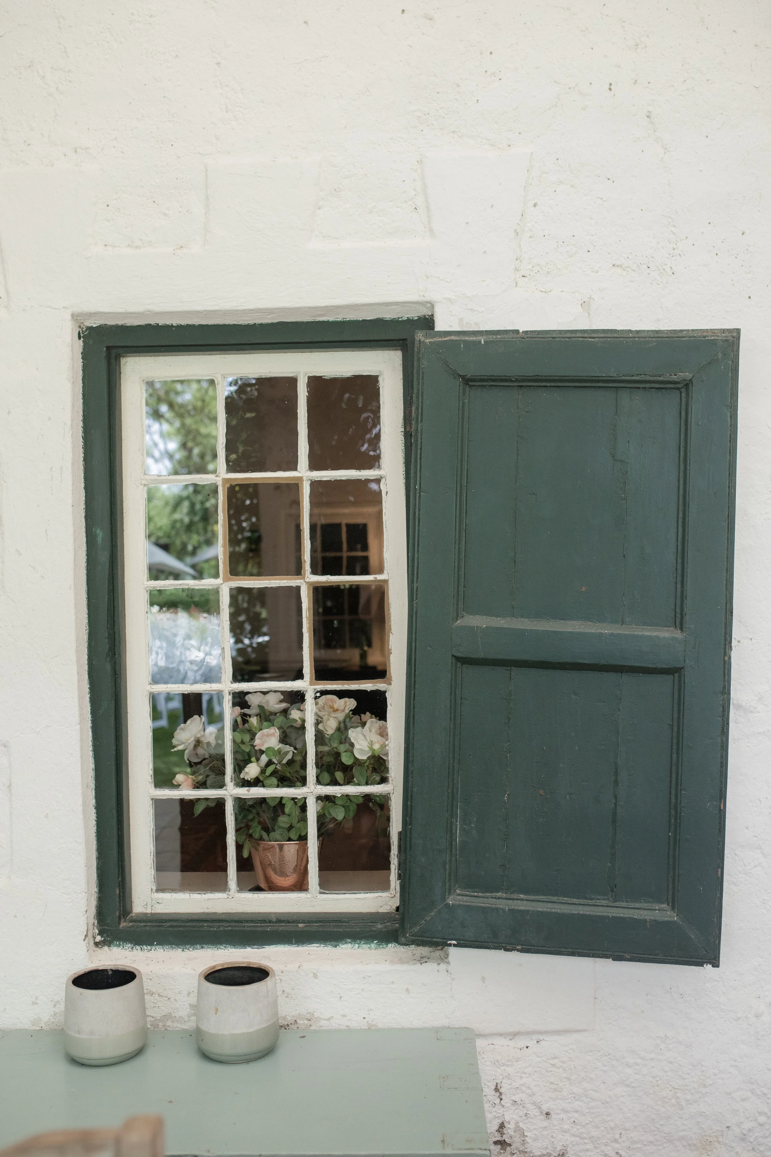 A white wall with a small window and a dark green wooden shutter, a potted plant with white flowers inside the window, and two white ceramic pots on a light green table.