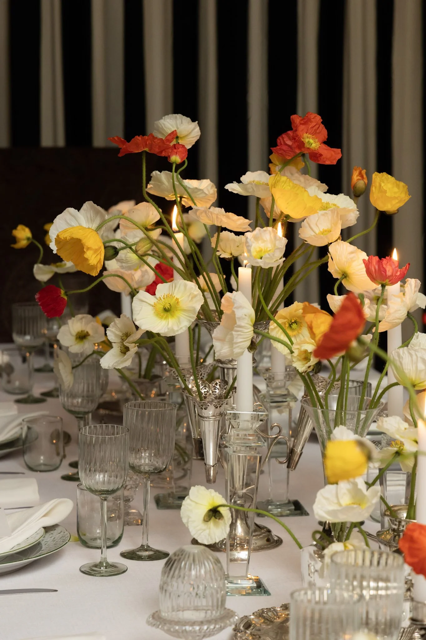 Elegant banquet table with tall floral arrangements of poppies in glass vases, surrounded by lit candles, glassware, and dinnerware on a white tablecloth, with dark curtains in the background.