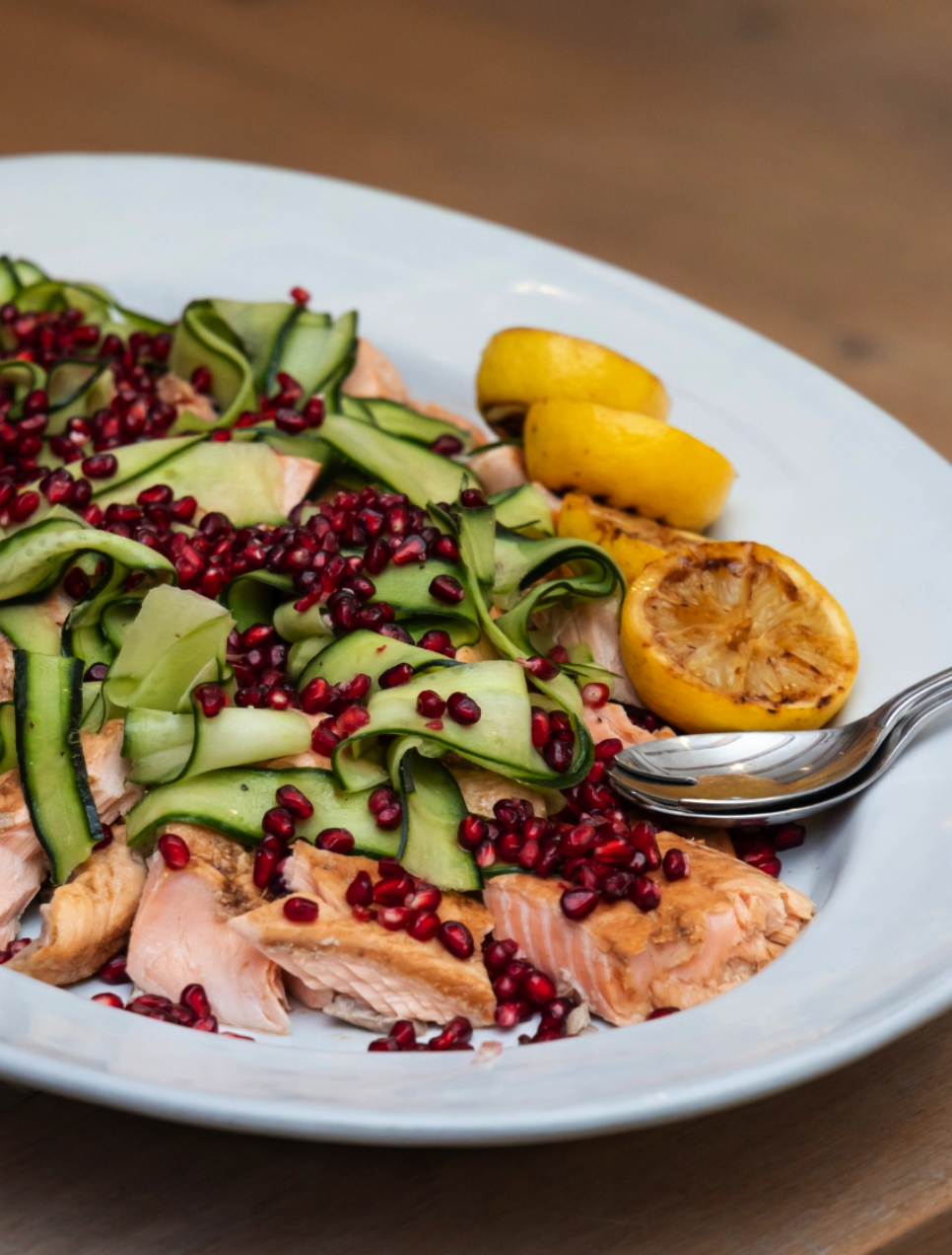 Salmon fish topped with sliced cucumbers, pomegranate seeds, and lemon wedges on a white plate.