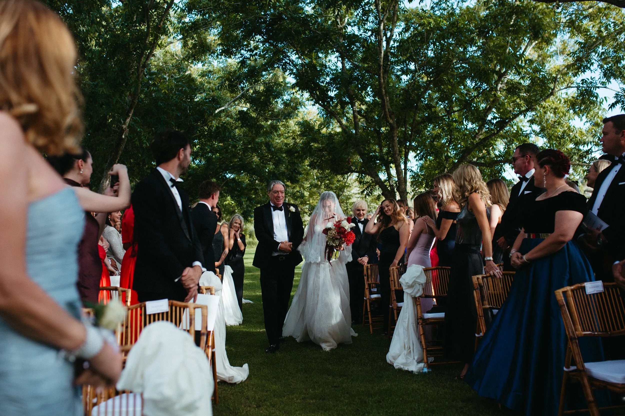 People gathered outdoors for a wedding ceremony, with a bride in a white gown holding a bouquet of red flowers walking down the aisle, and guests seated and standing around under tall trees.