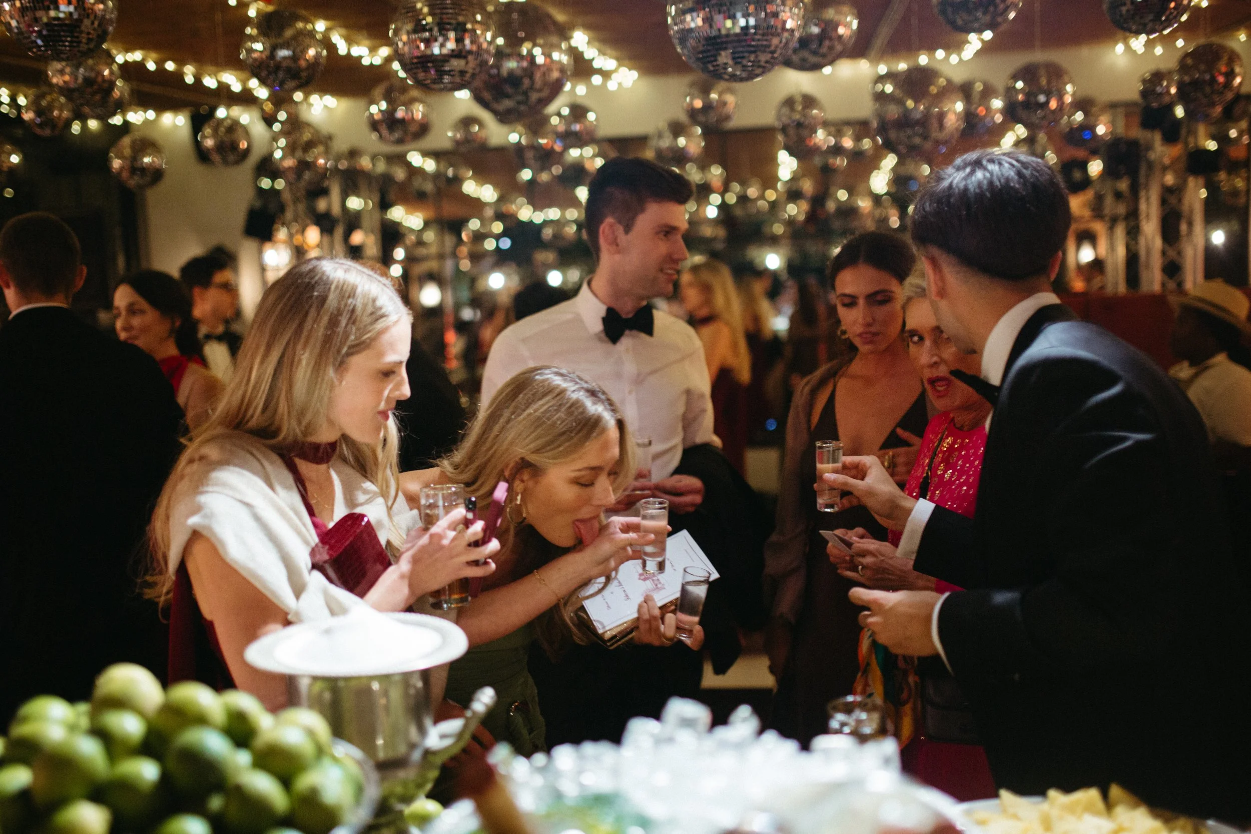 People celebrating at a festive gathering with disco balls and string lights, with a table of food and drinks in the foreground.