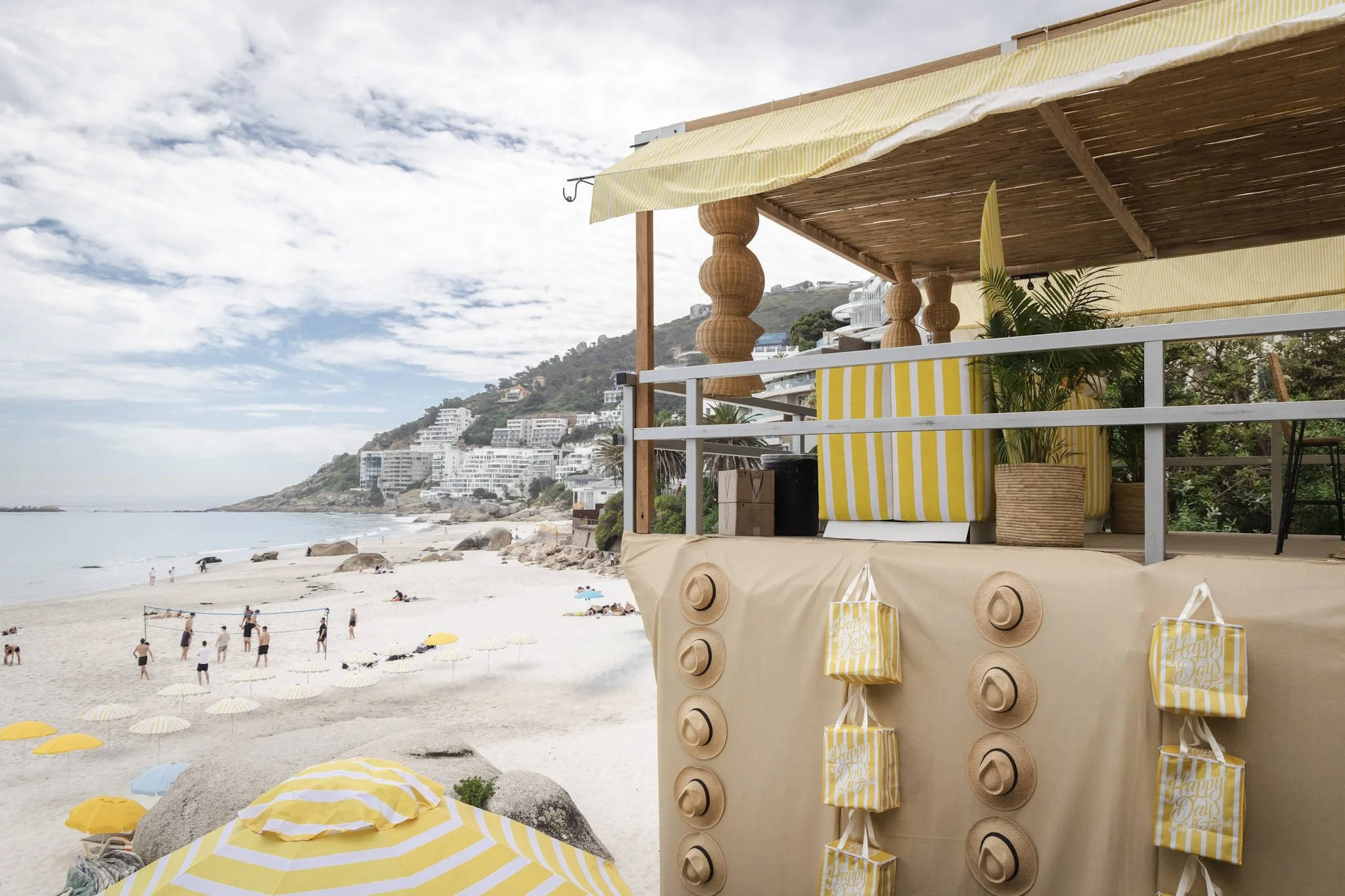 Beachside cabana with yellow and white striped awning, yellow and white striped bags, and a potted plant, overlooking a beach with umbrellas, rocks, and people, with buildings on a hillside under a partly cloudy sky.