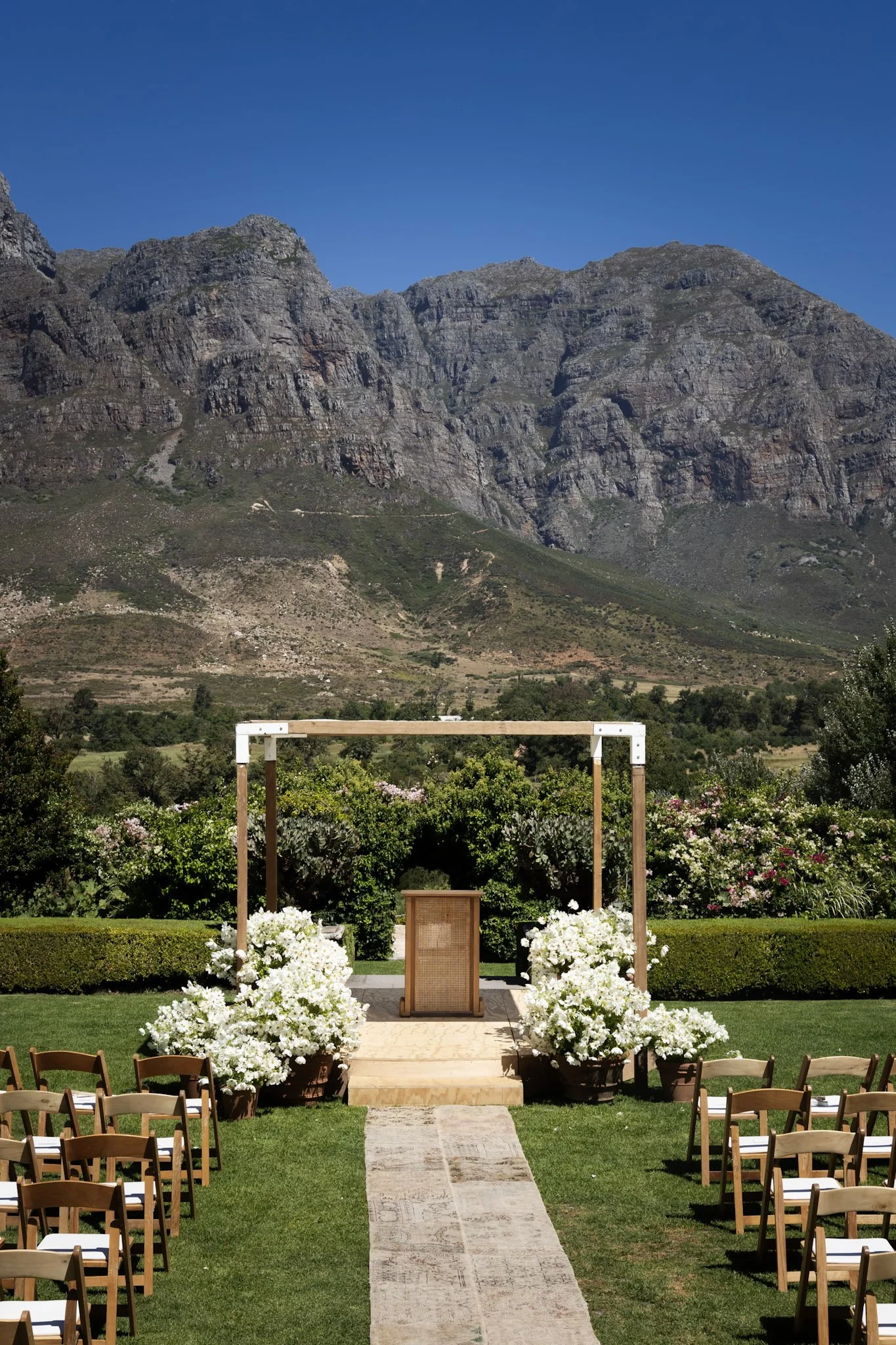 Outdoor wedding ceremony setup with chairs arranged on either side of an aisle, white flowers, and a wooden arch, set against a backdrop of mountains and a blue sky.
