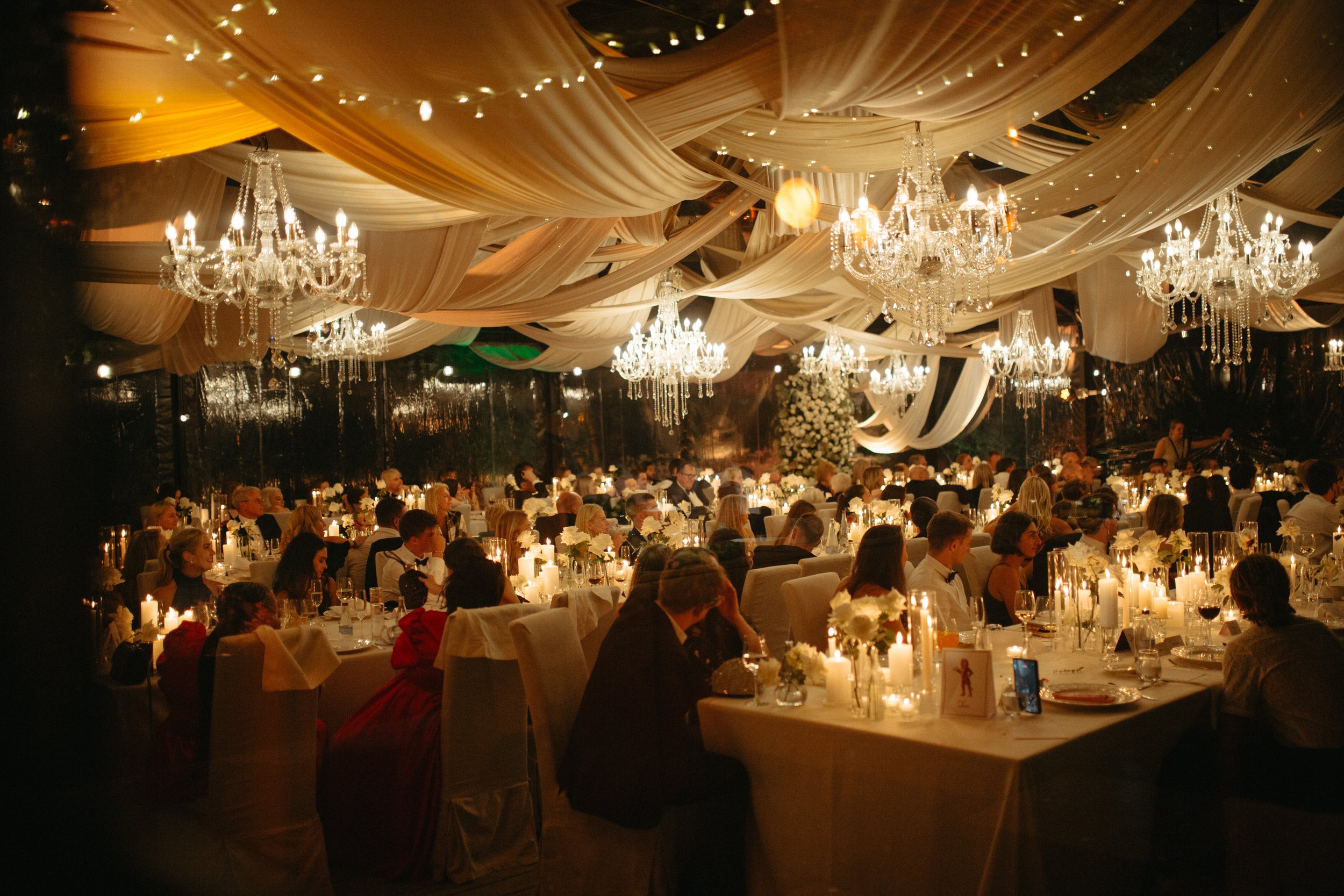 A large banquet hall decorated with draped fabric ceiling, chandeliers, and warm lighting. Long tables are set for a formal dinner with white flowers and lit candles. Many guests are seated, dressed in formal attire, enjoying the event.