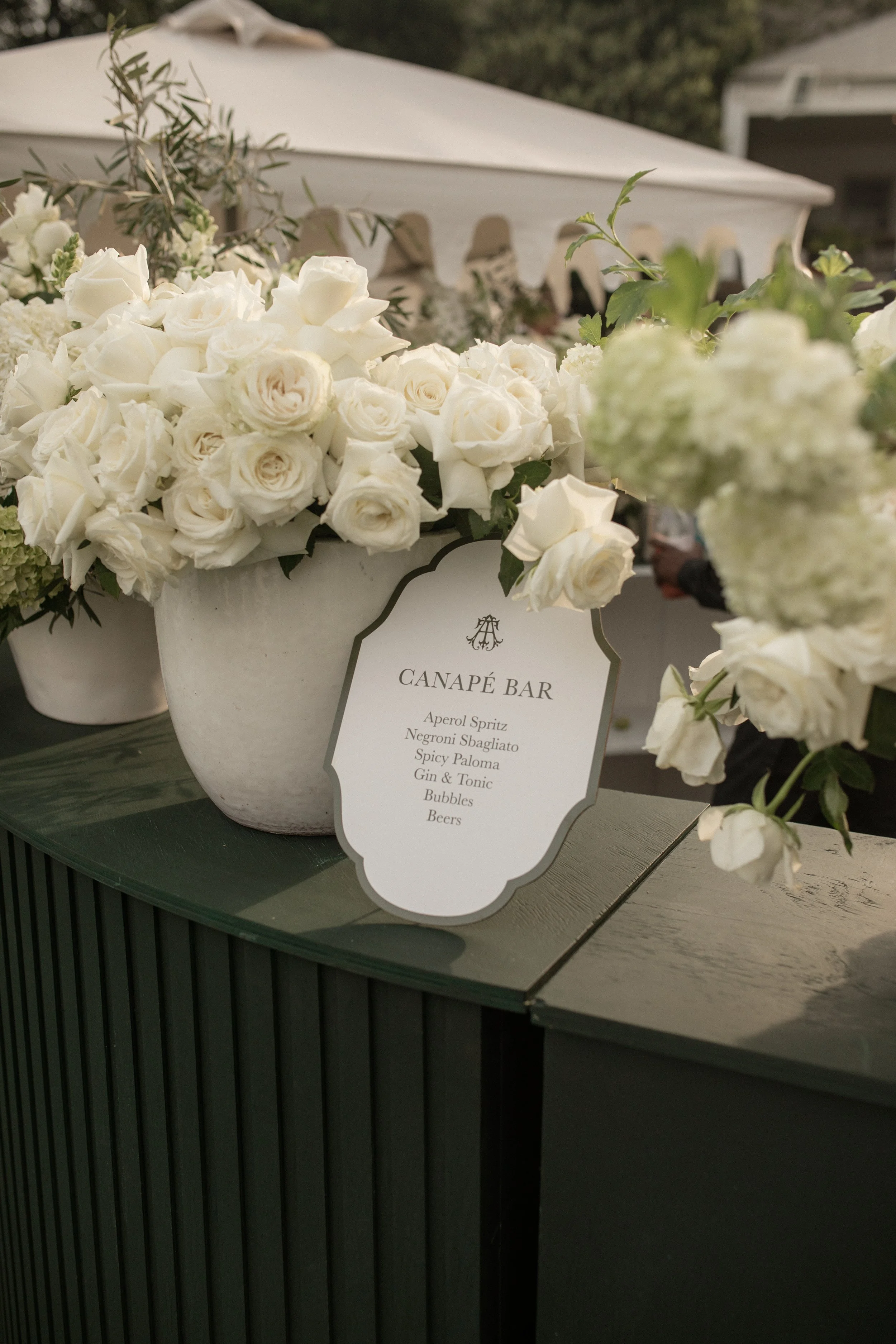 A white flower arrangement on a table with a sign labeled "Canapé Bar" listing various drinks, surrounded by greenery and a white canopy in the background.
