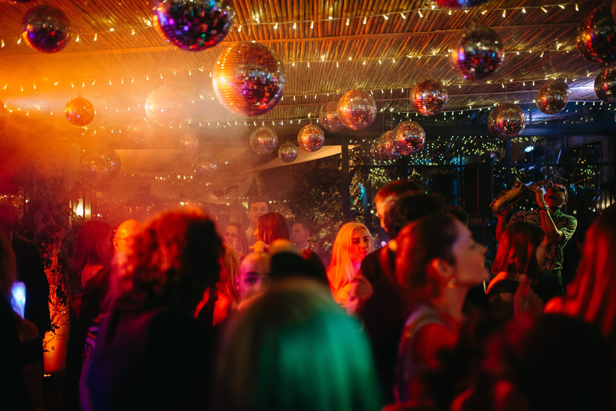 People dancing and socializing at a party with disco balls, string lights, and colorful lighting.